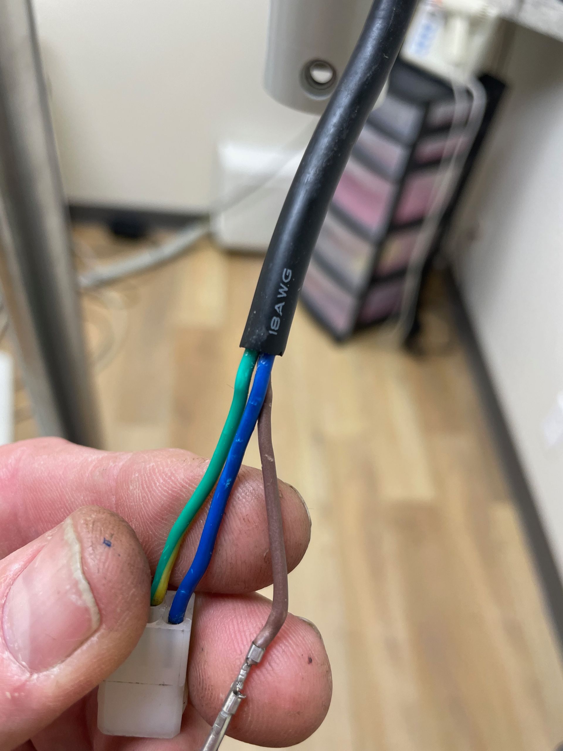 Hand holding a stripped cable with green, blue, and brown wires, attached to a white connector. A black cable is visible.