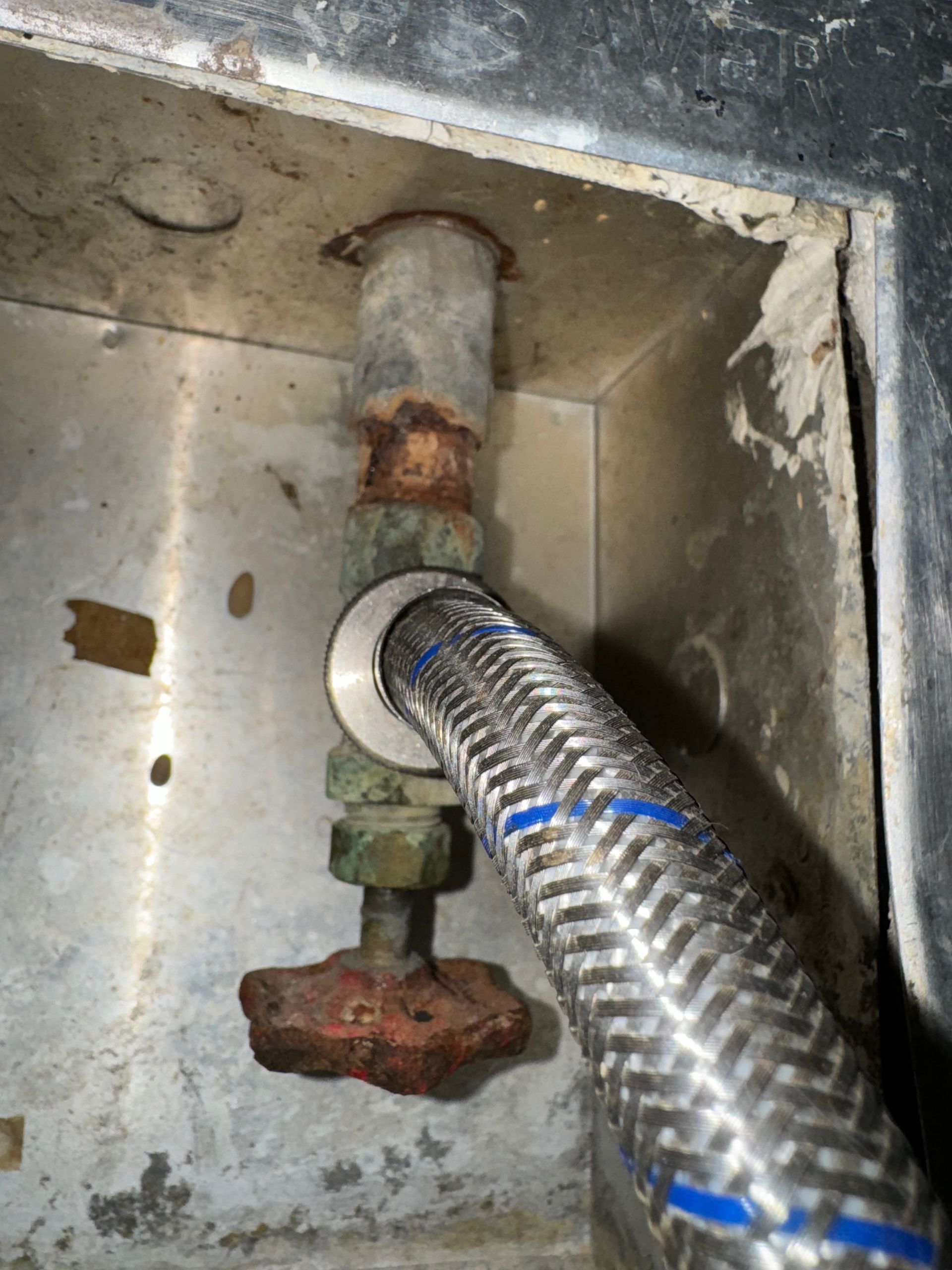 A plumbing setup with a corroded pipe and red valve, connected to a braided hose within a metal enclosure.