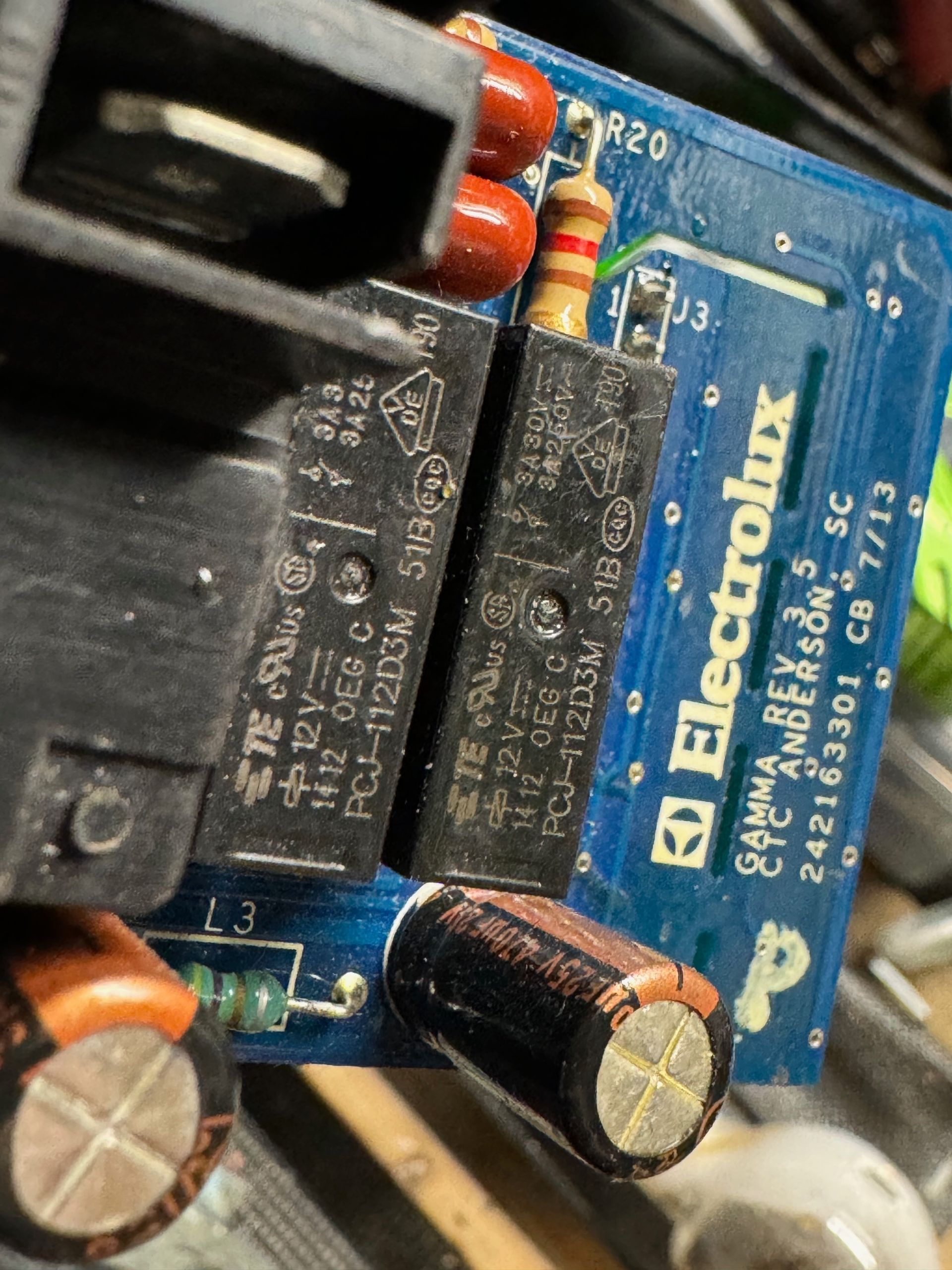 Close-up of a blue Electrolux circuit board with two black relays, brown and yellow resistors, and a black capacitor.