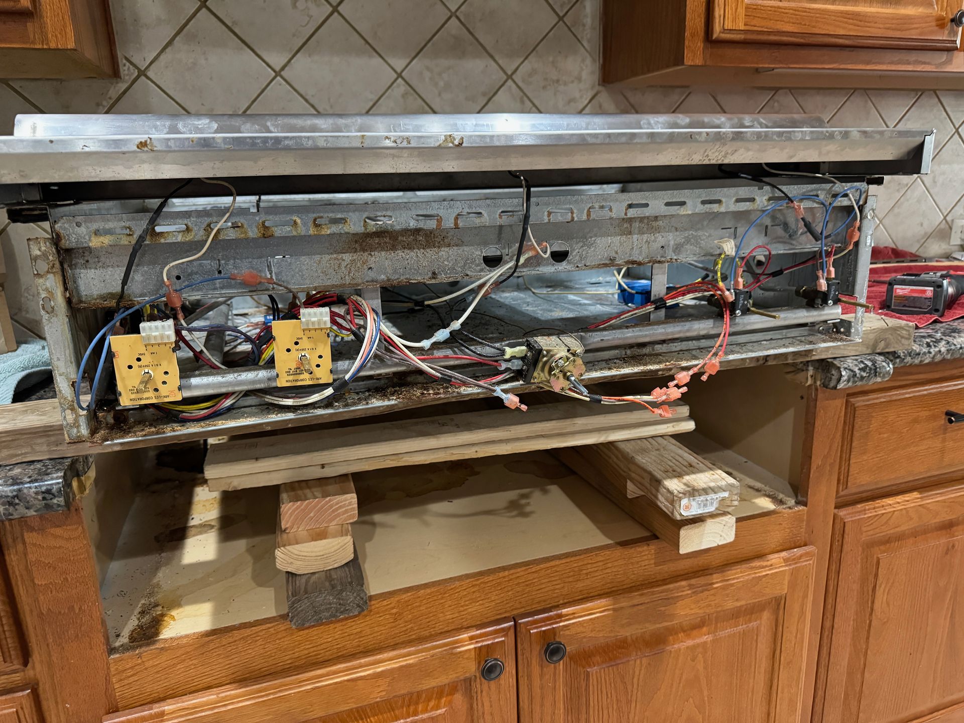 A disassembled stovetop rests on makeshift supports in a partially open kitchen cabinet. Wires and internal components are visible.