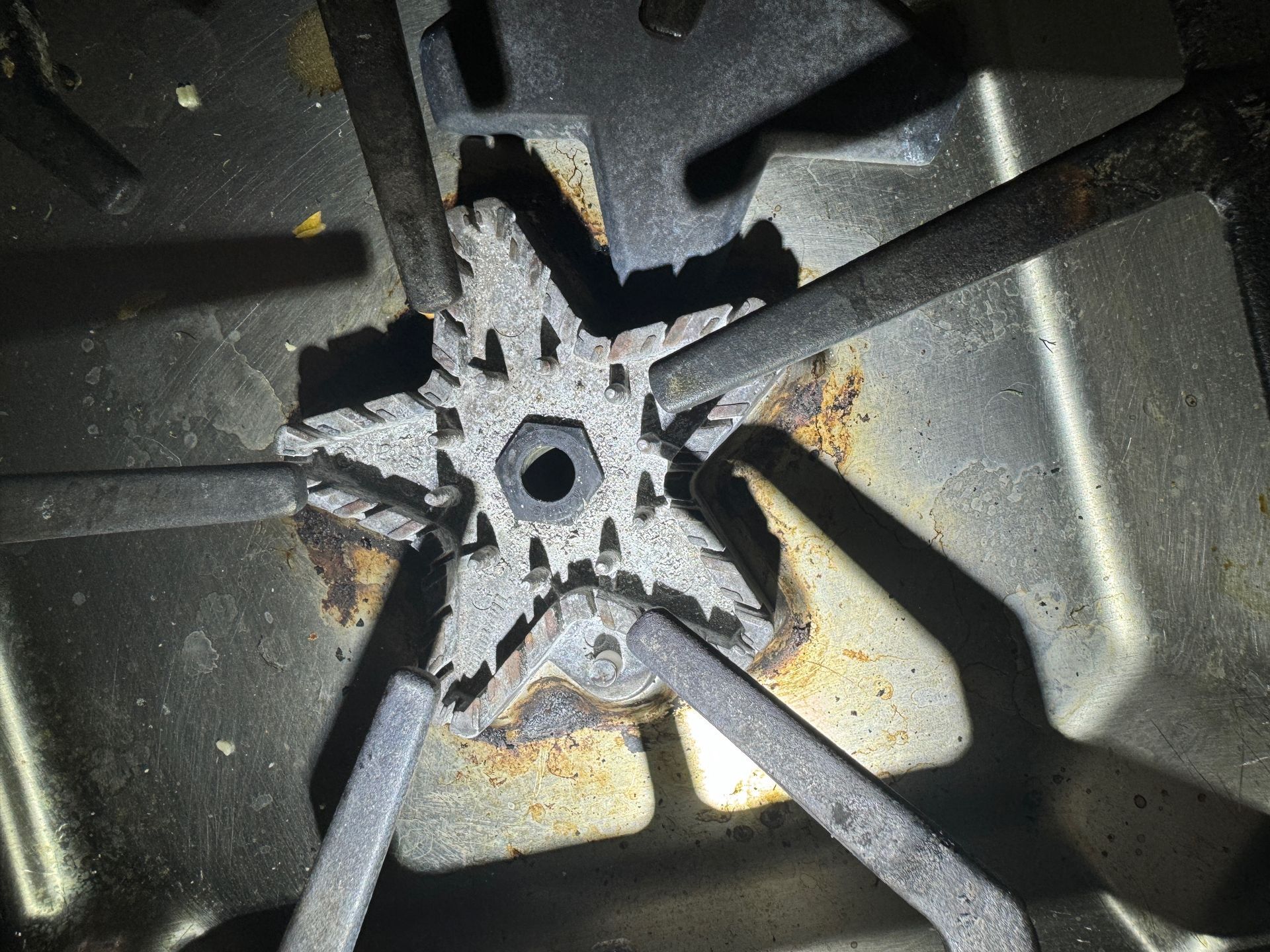 Close-up of a gas stovetop burner with a star-shaped burner head and radiating metal arms, showing signs of rust.
