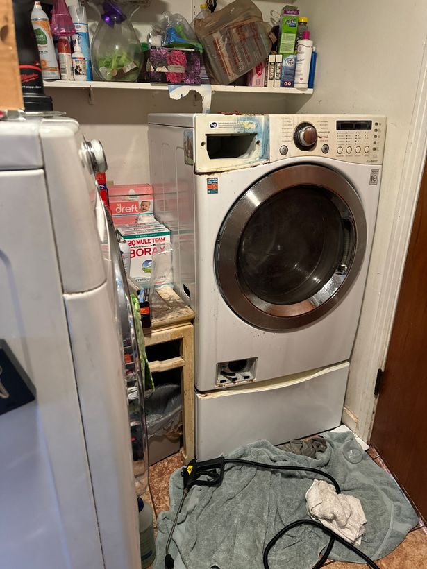 A white front-loading washing machine, possibly damaged, in a cluttered laundry area with cleaning supplies.