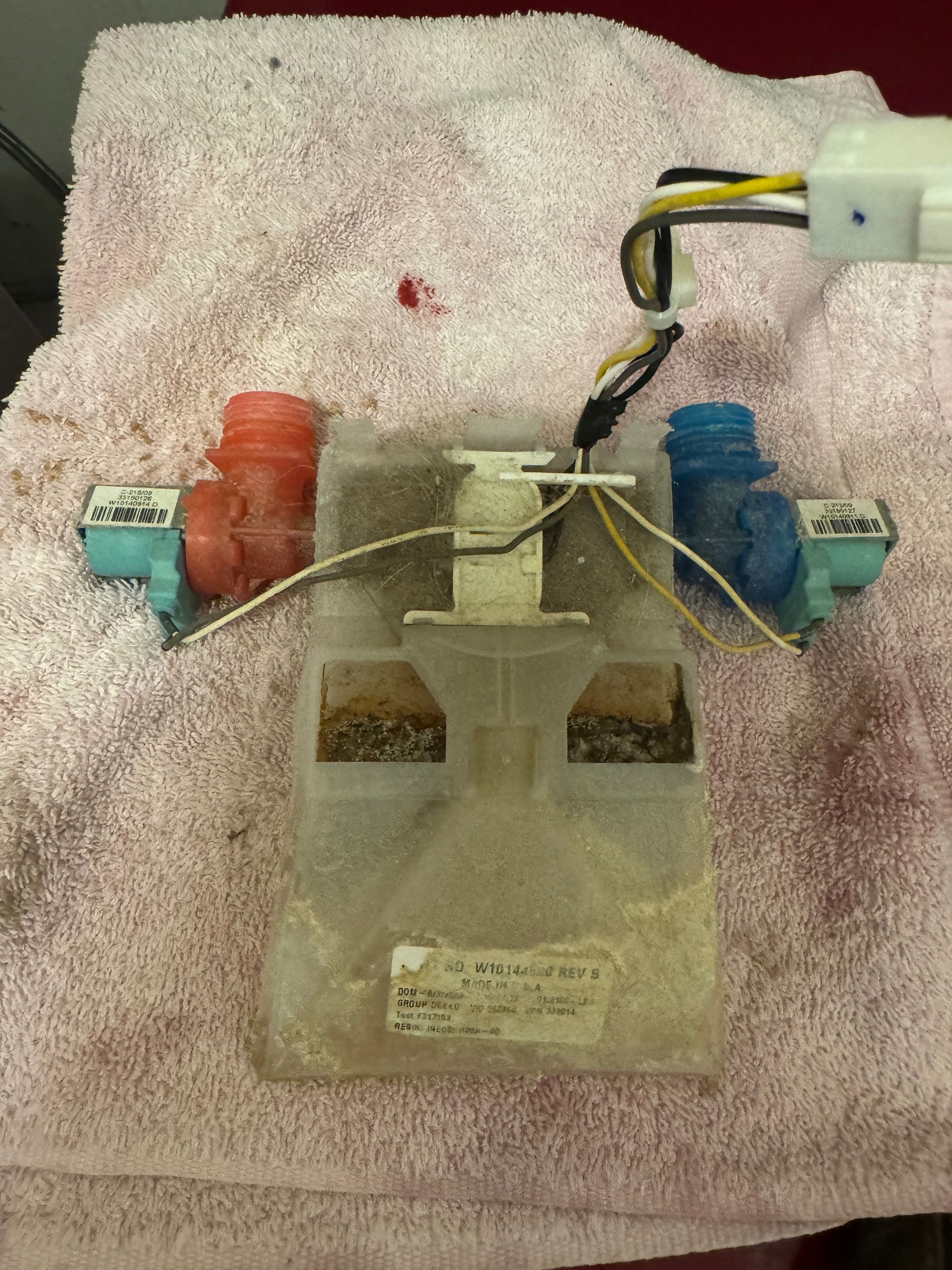 A washing machine water inlet valve assembly with blue and red plastic connectors, a white plastic body, and electrical wiring on a pink towel.