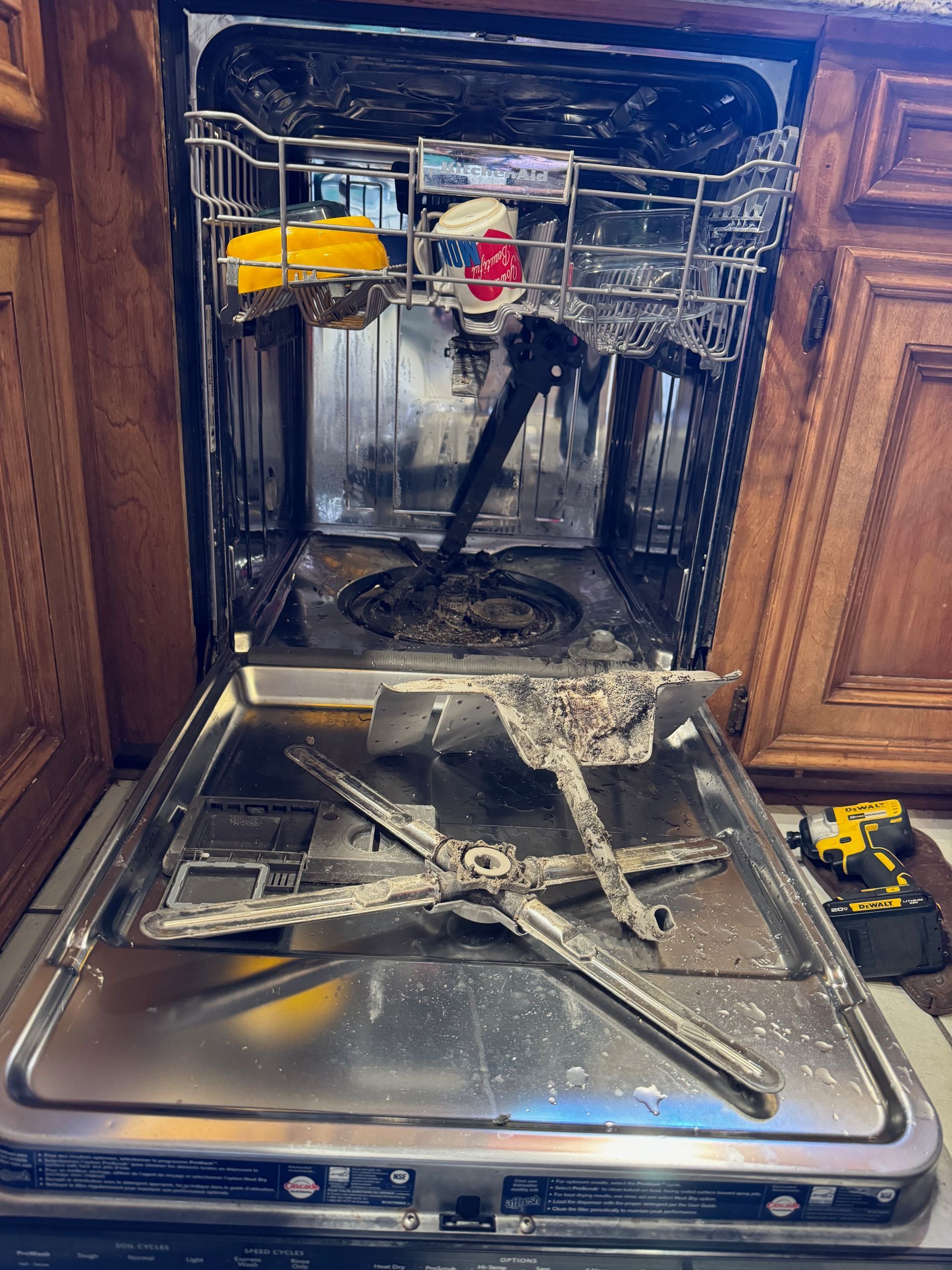 Open dishwasher with debris and a broken spray arm. Interior appears dirty, with wooden cabinets surrounding the appliance.