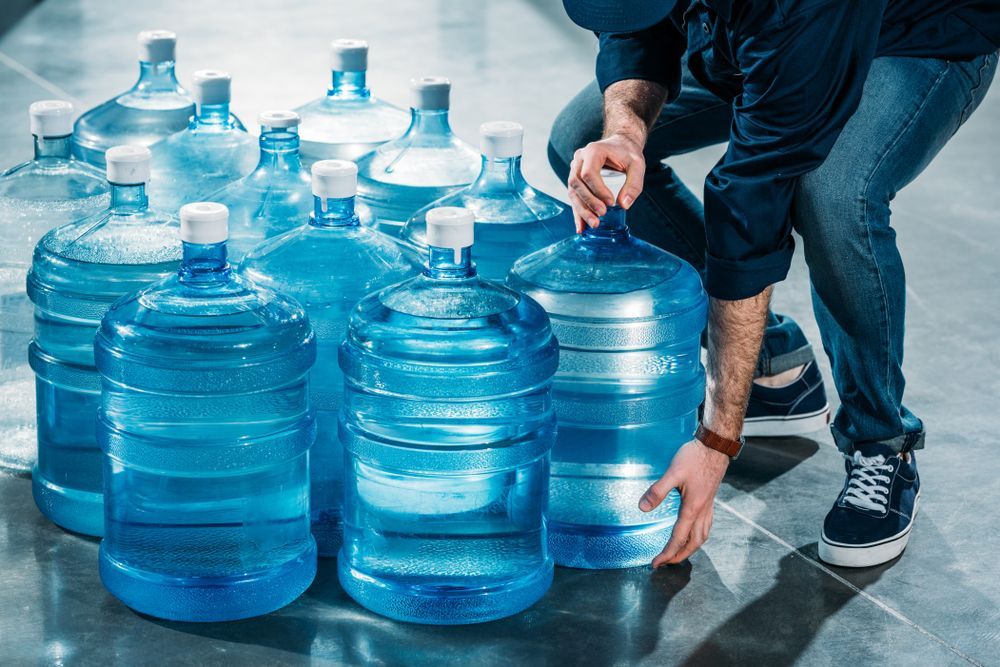 Person Arranging Large Blue Water Jugs on a Tiled Floor in a Storage Area — Ts Jeffree Contractors in Tennant Creek, QLD