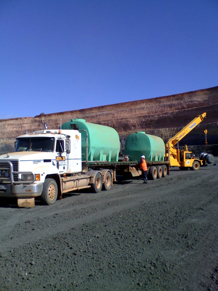 Truck carrying two large green water tanks on a gravel road, with a forklift and person in an orange vest— Ts Jeffree Contractors in Burketown, QLD