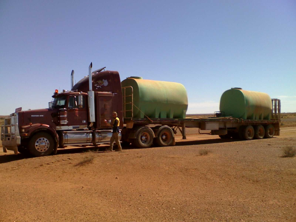 Red semi-truck hauling two green water tanks on a desolate dirt road under a blue sky.— Ts Jeffree Contractors in Burketown, QLD