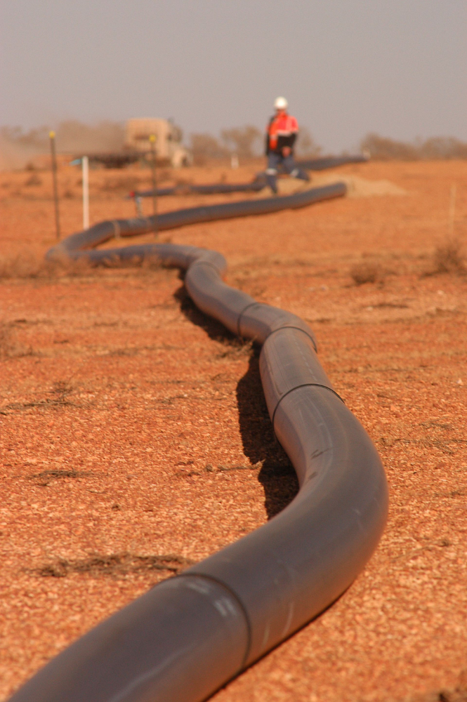 Black pipeline snaking across red dirt, worker in distance.— Ts Jeffree Contractors in Mount Isa, QLD