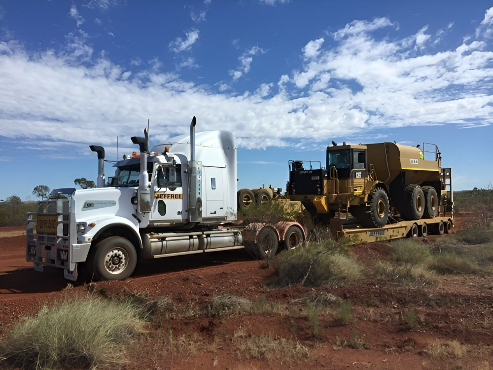 White semi-truck transporting yellow construction vehicle on a flatbed trailer, outdoors on red dirt under a blue sky.— Ts Jeffree Contractors in Normanton, QLD