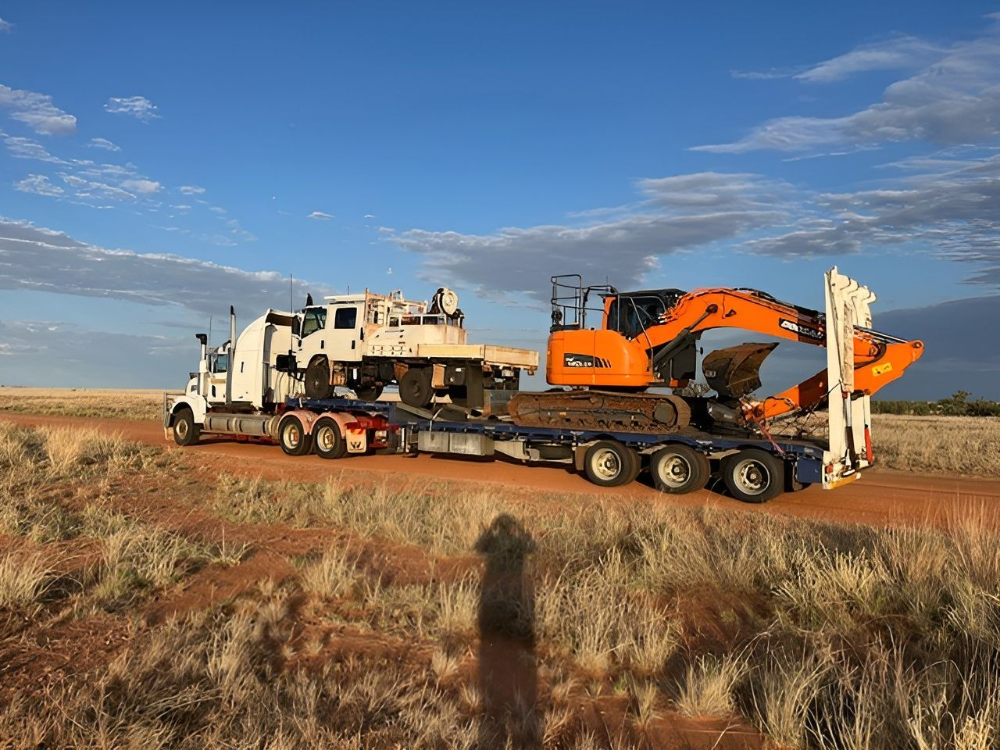 Truck hauling an excavator and a large white structure on a lowboy trailer on a dirt road. Blue sky— Ts Jeffree Contractors in Normanton, QLD