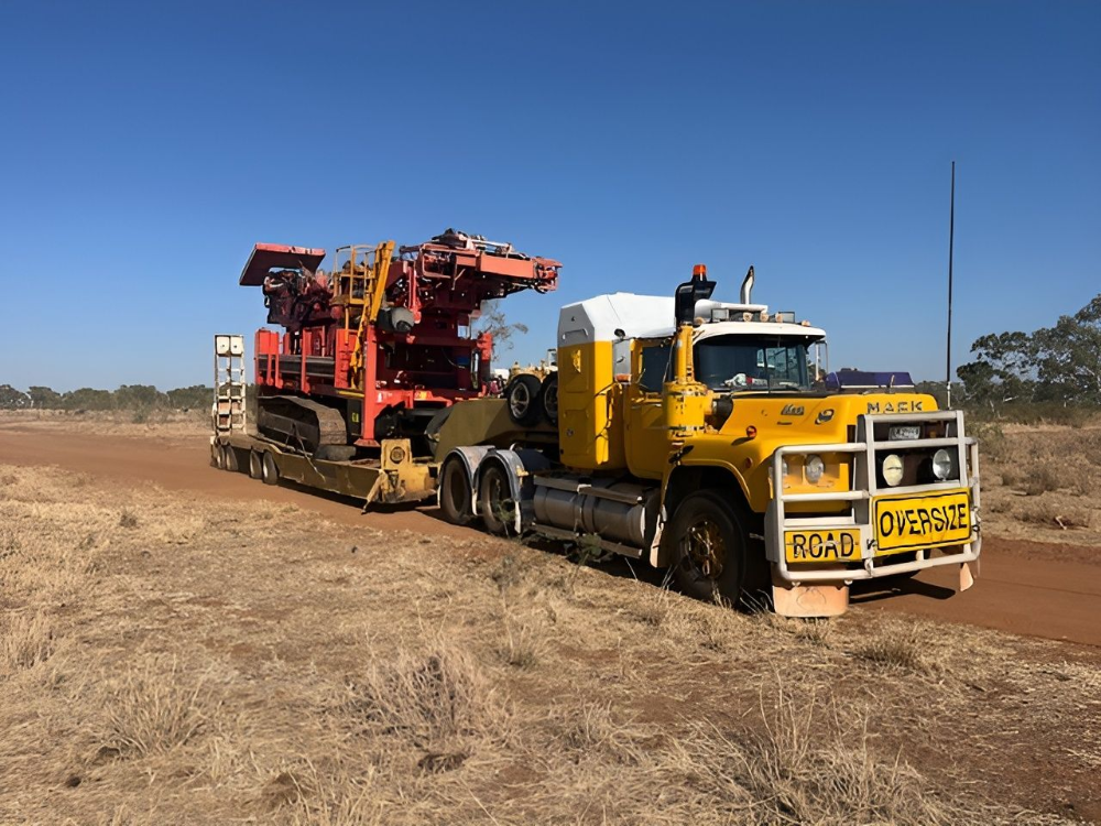 Yellow truck transporting a large red drilling machine on a low-loader trailer on a dirt road.— Ts Jeffree Contractors in Tennant Creek, NT