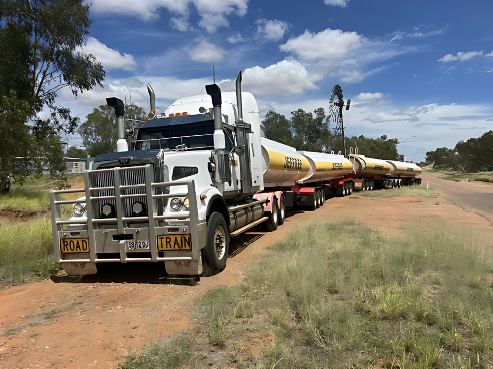 White semi-truck hauling long yellow cylindrical pipes on a dirt road under a blue sky