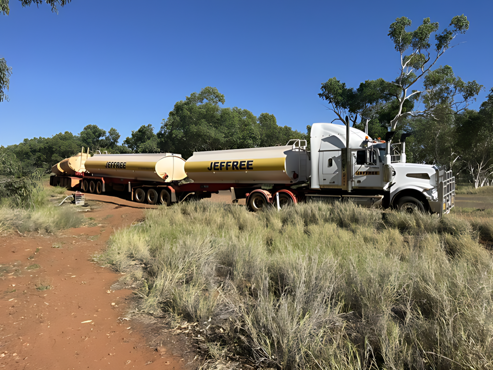 White tanker truck hauling two long cream tanks on a dirt road in a dry, grassy area under a blue sky— Ts Jeffree Contractors in Cloncurry, QLD