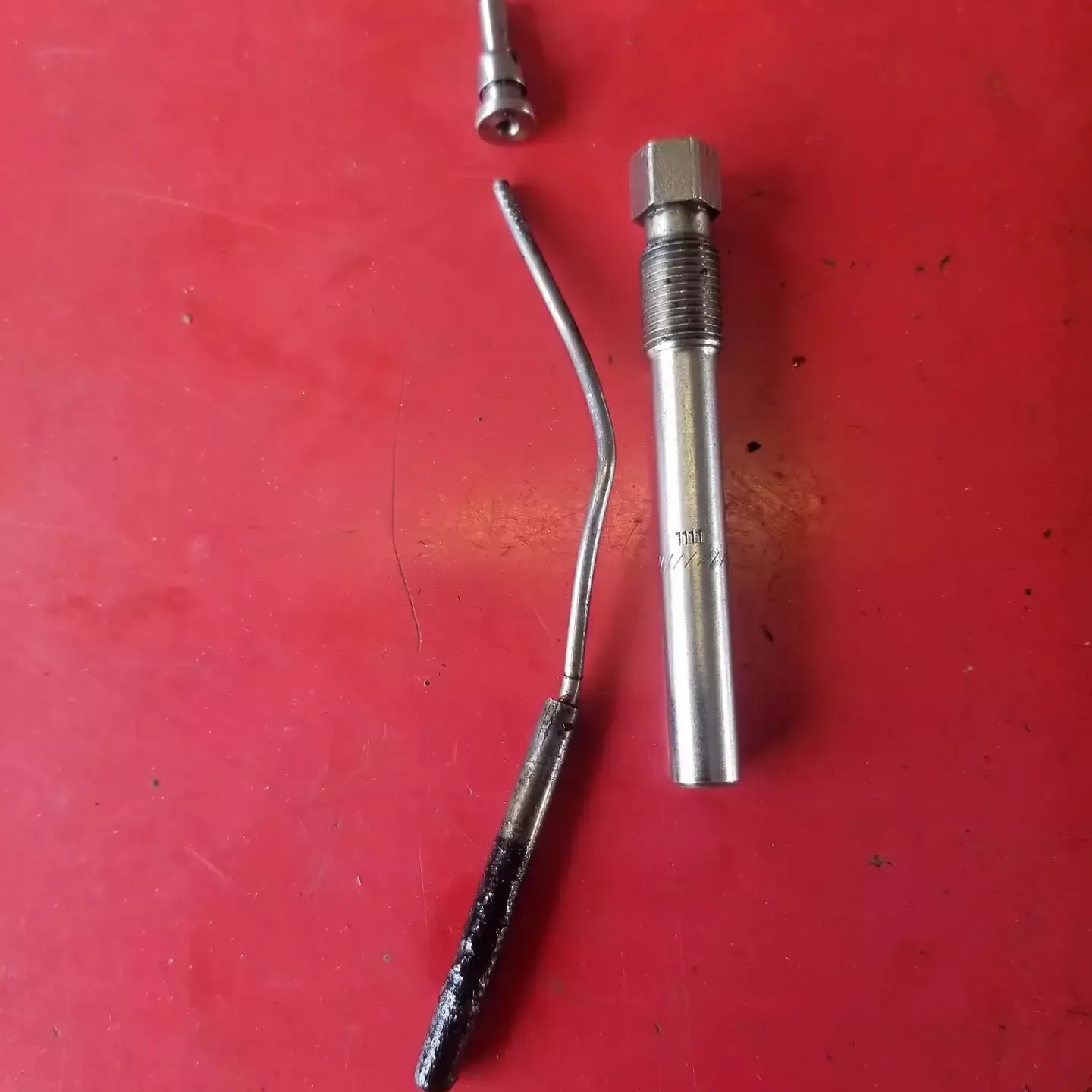 Parts laid out on a red surface: a long metallic tube, a curved metal rod, and a smaller bolt.