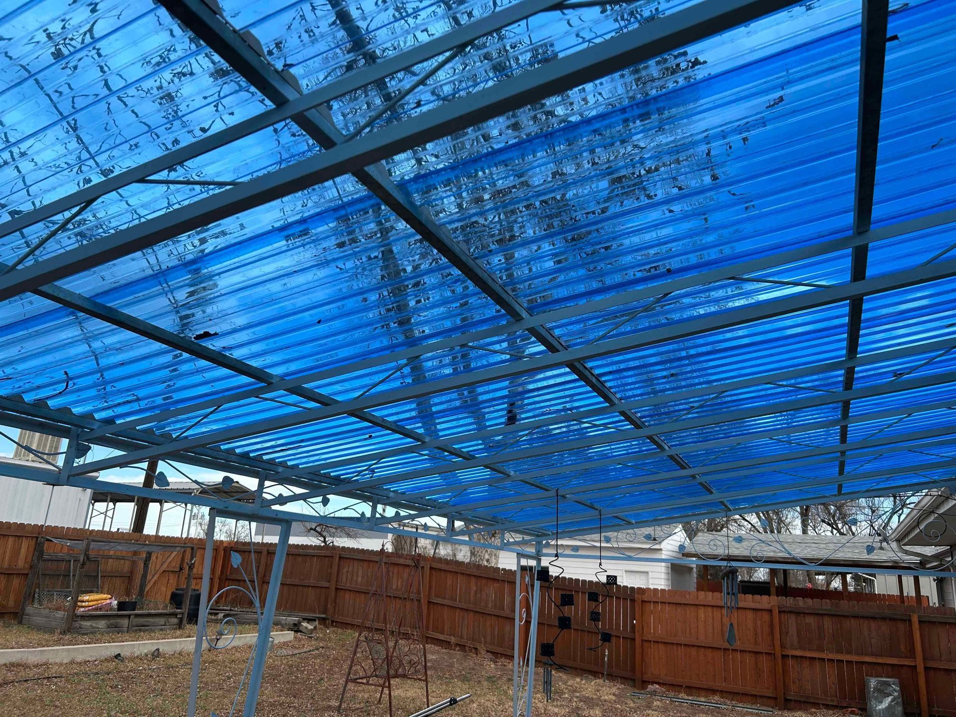 Blue corrugated roof over metal frame, covering a backyard patio. A brown fence surrounds the yard.