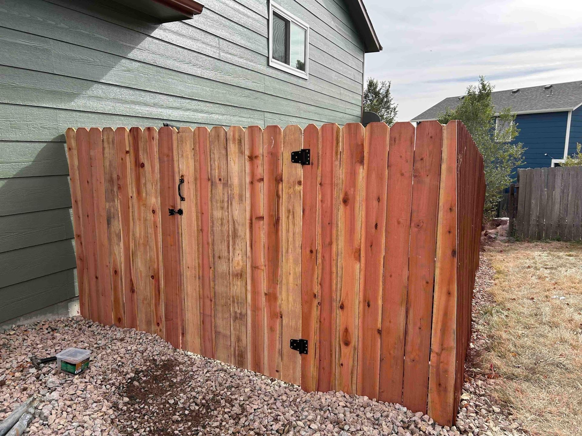Wooden fence attached to a green house, with a gate and gravel ground.