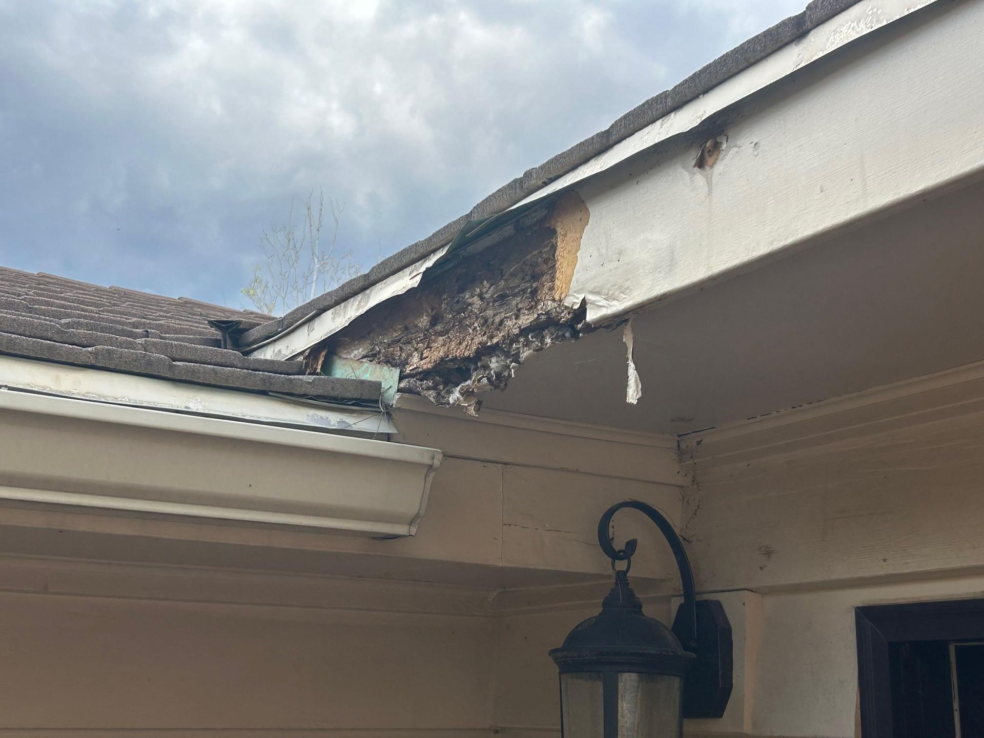 Damaged roof edge with exposed wood. Brown shingles, white trim, and a black outdoor light. Cloudy sky.