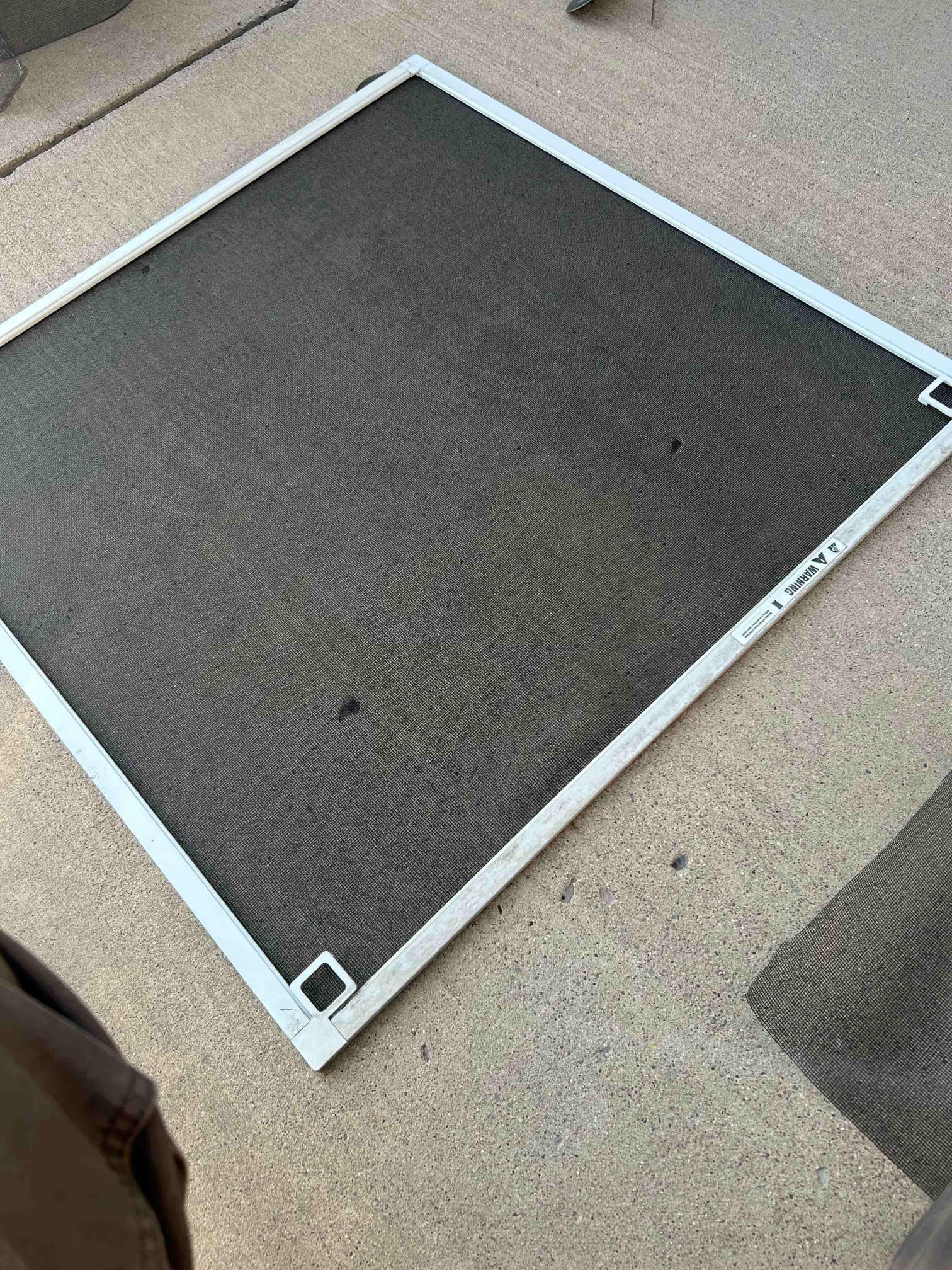 White-framed window screen with dark mesh, lying on a gray concrete surface.