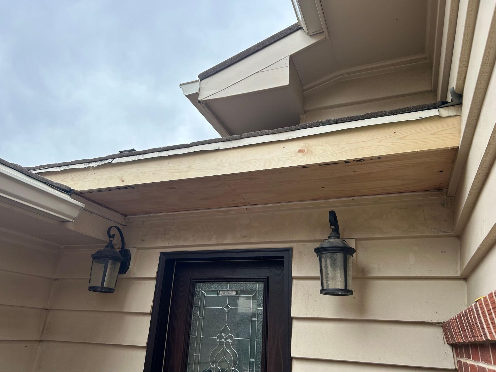Exterior view of a house entry with a door, lights, and exposed wooden framing under the roof's overhang.