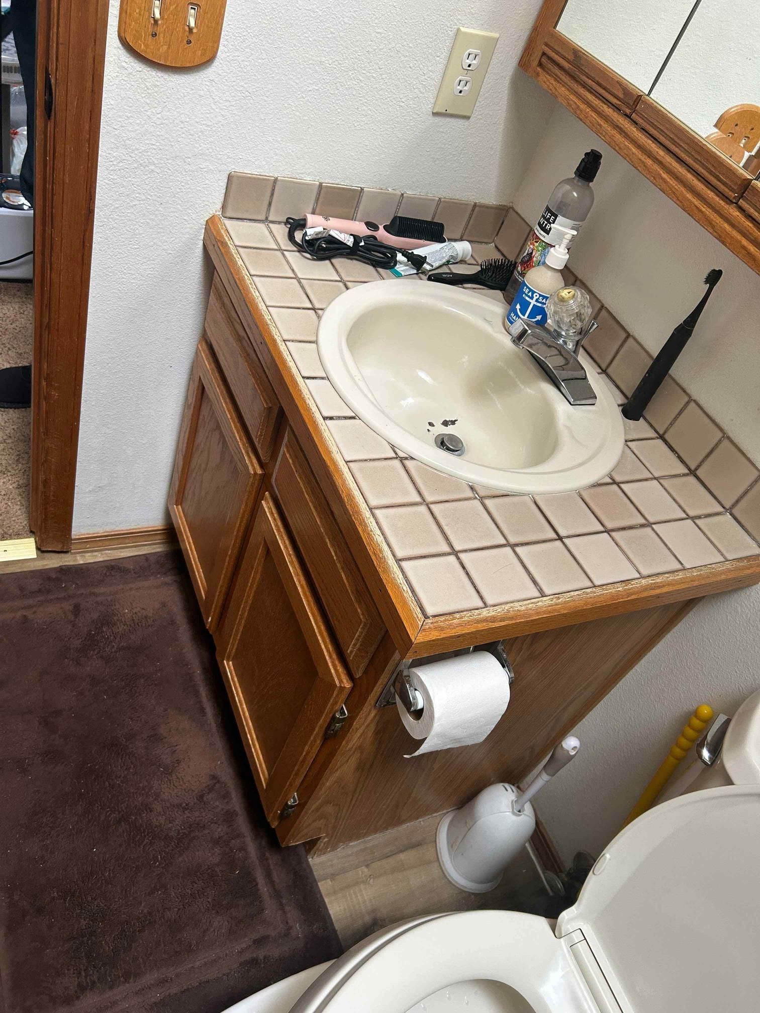 Bathroom vanity with sink, cabinet, and tiled countertop. Toilet paper roll and toilet are visible.