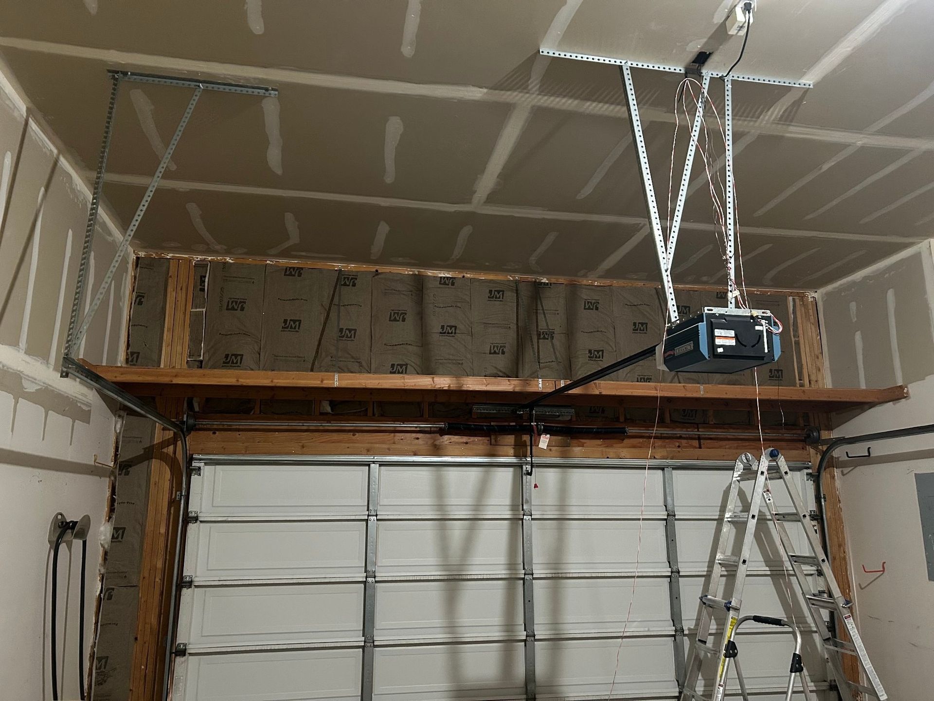Garage interior with exposed framing, insulation, and drywall. Garage door is partially visible. A ladder is present.