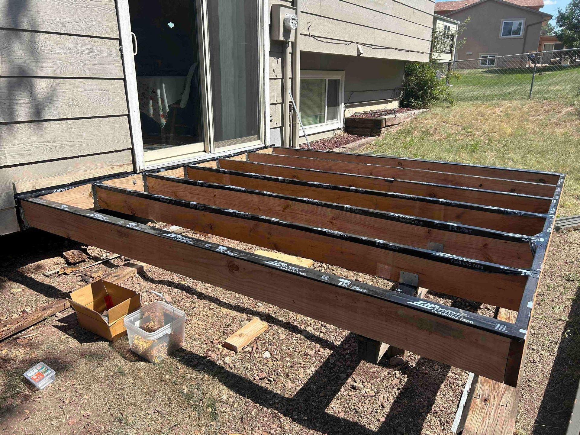 Wooden deck frame under construction outside a house with a sliding glass door.