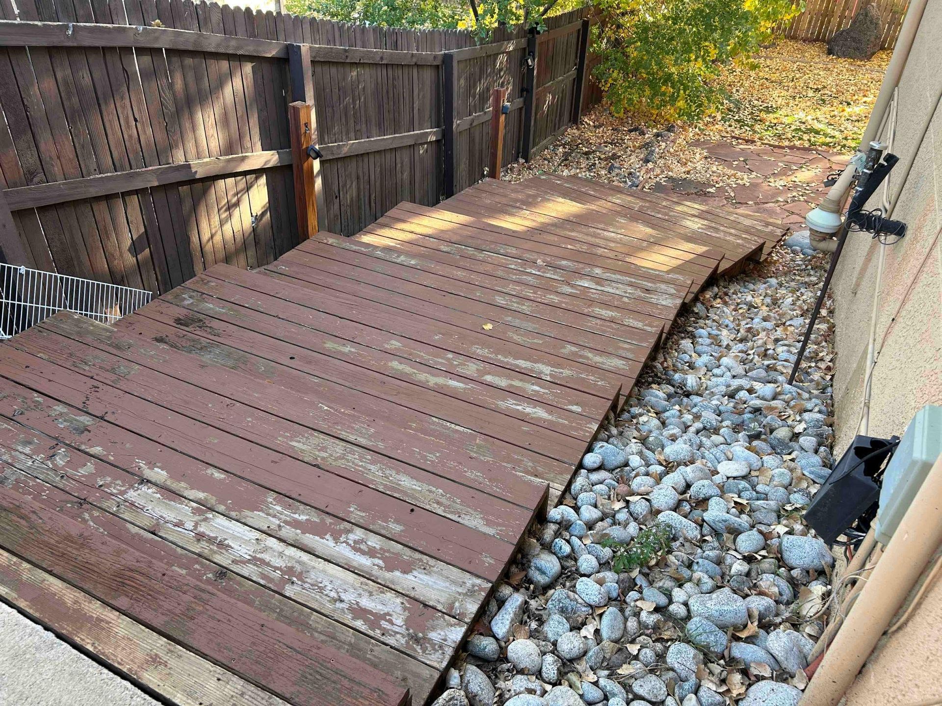 Dilapidated wooden walkway alongside a building and gravel. Brown fence in background.