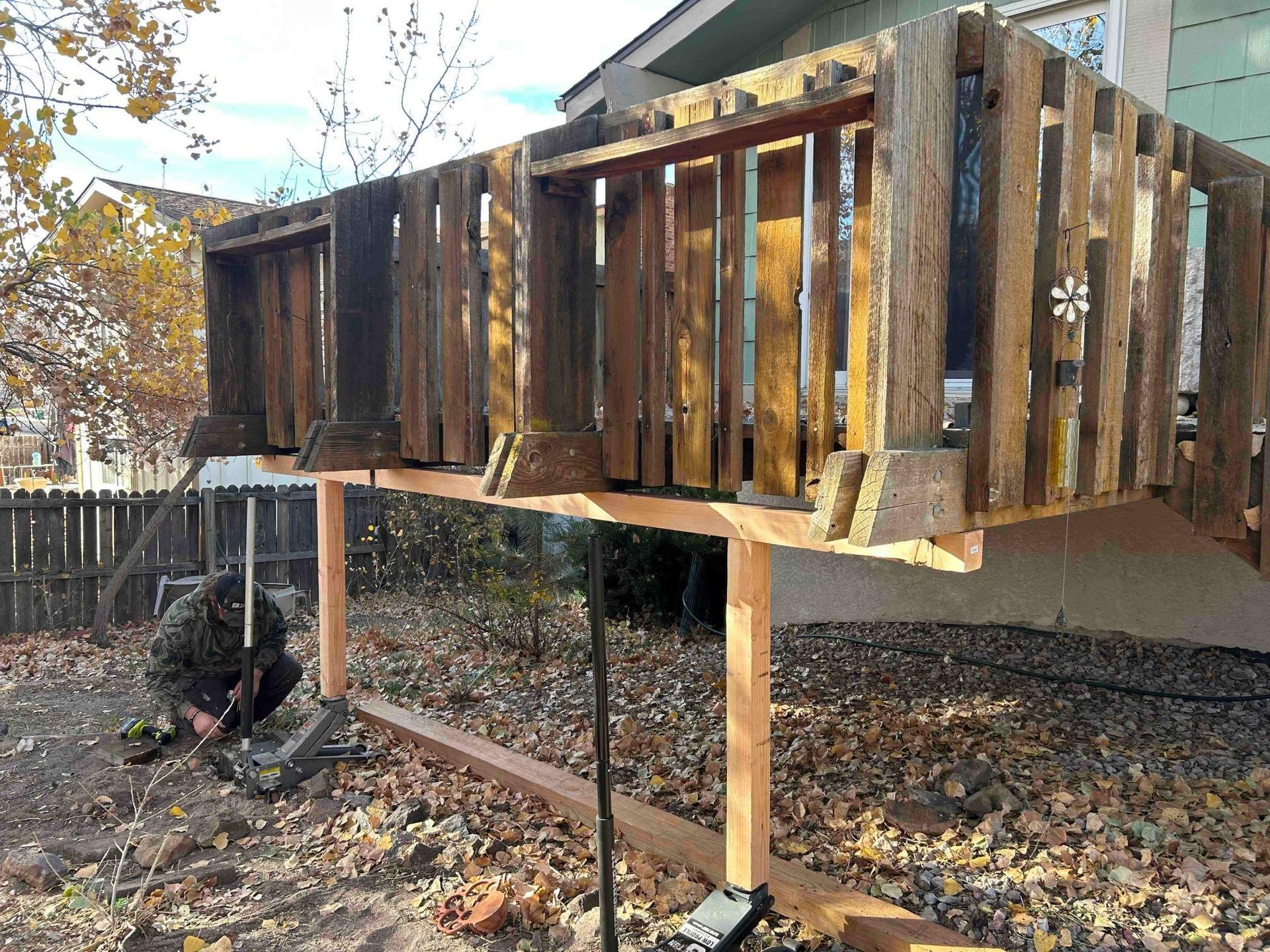 Person working on a wooden deck supported by wooden posts near a house and a fence, outside on a fall day.