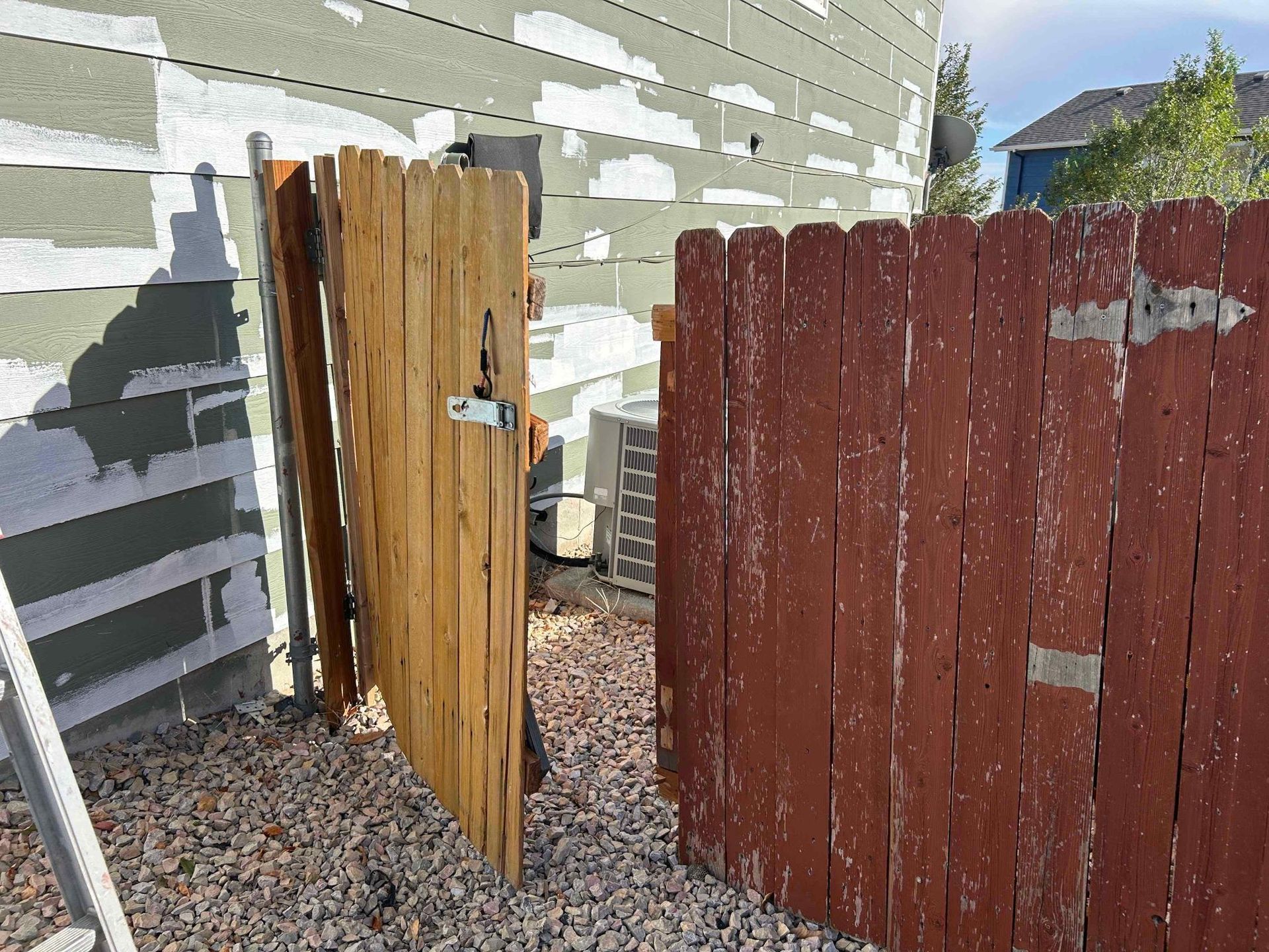 Two wooden gates, one tan and one red, next to a building with peeling stucco and an air conditioning unit.