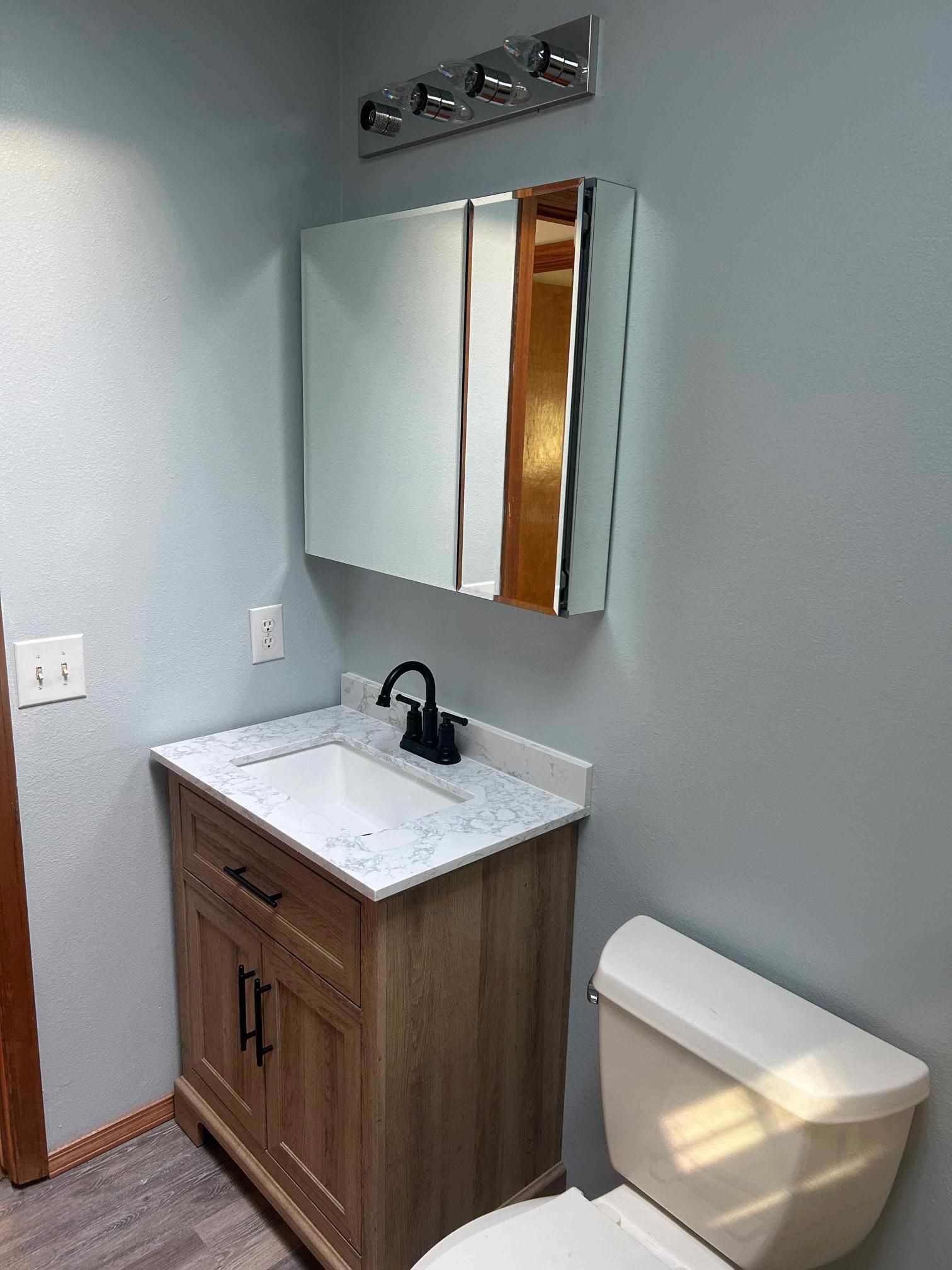 Bathroom with light blue walls, wood-look vanity, marble-like countertop, black faucet, and mirrored cabinet.