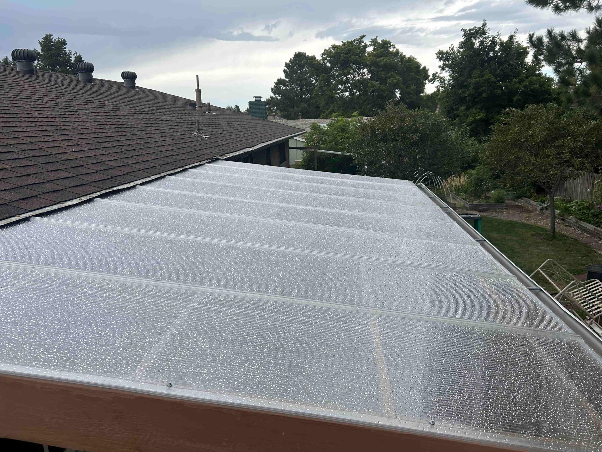 Clear, corrugated plastic roof over a deck, next to a brown shingled roof with trees in the background.
