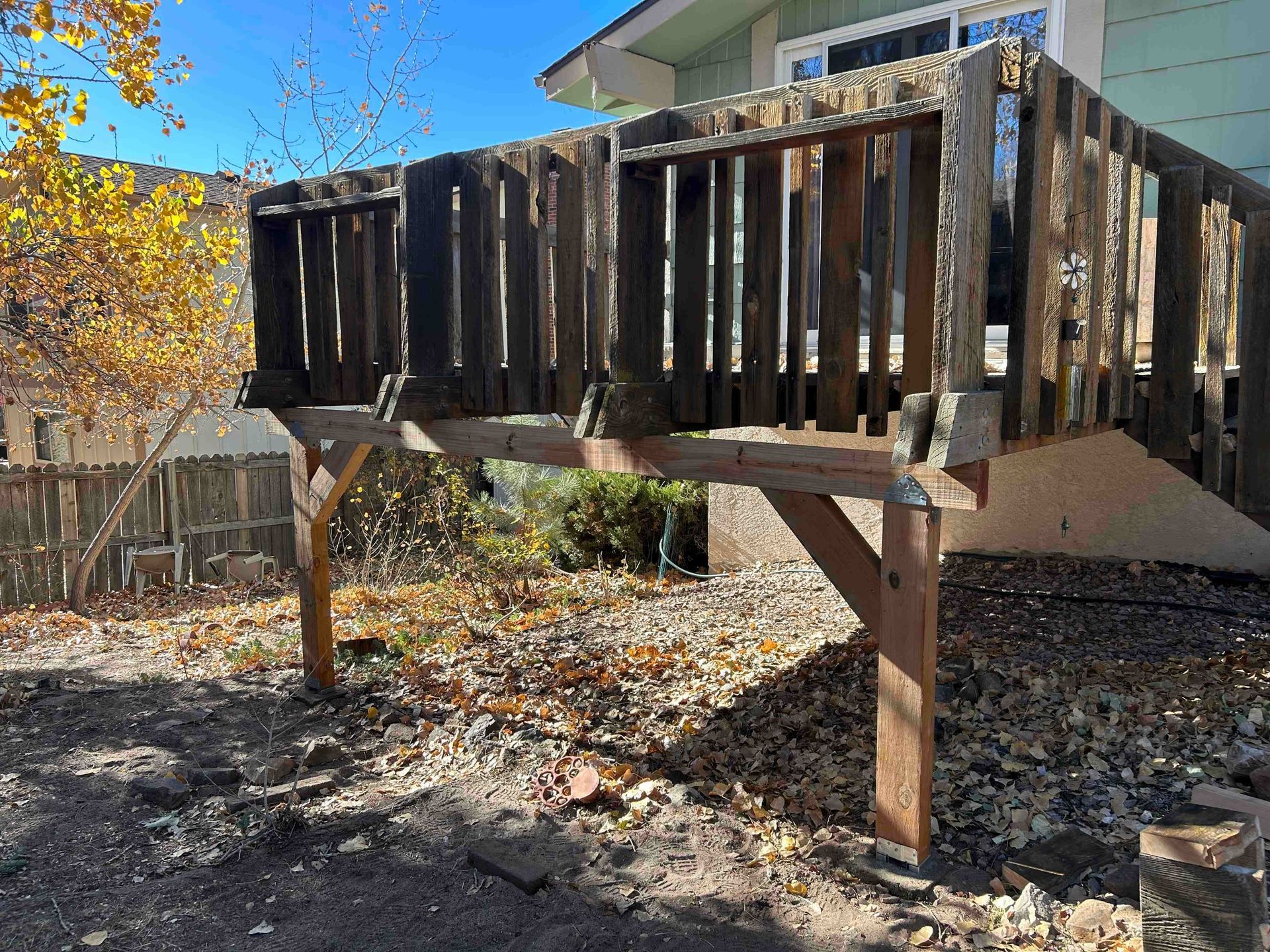 A weathered wooden deck with a railing stands on posts in a yard covered in leaves.