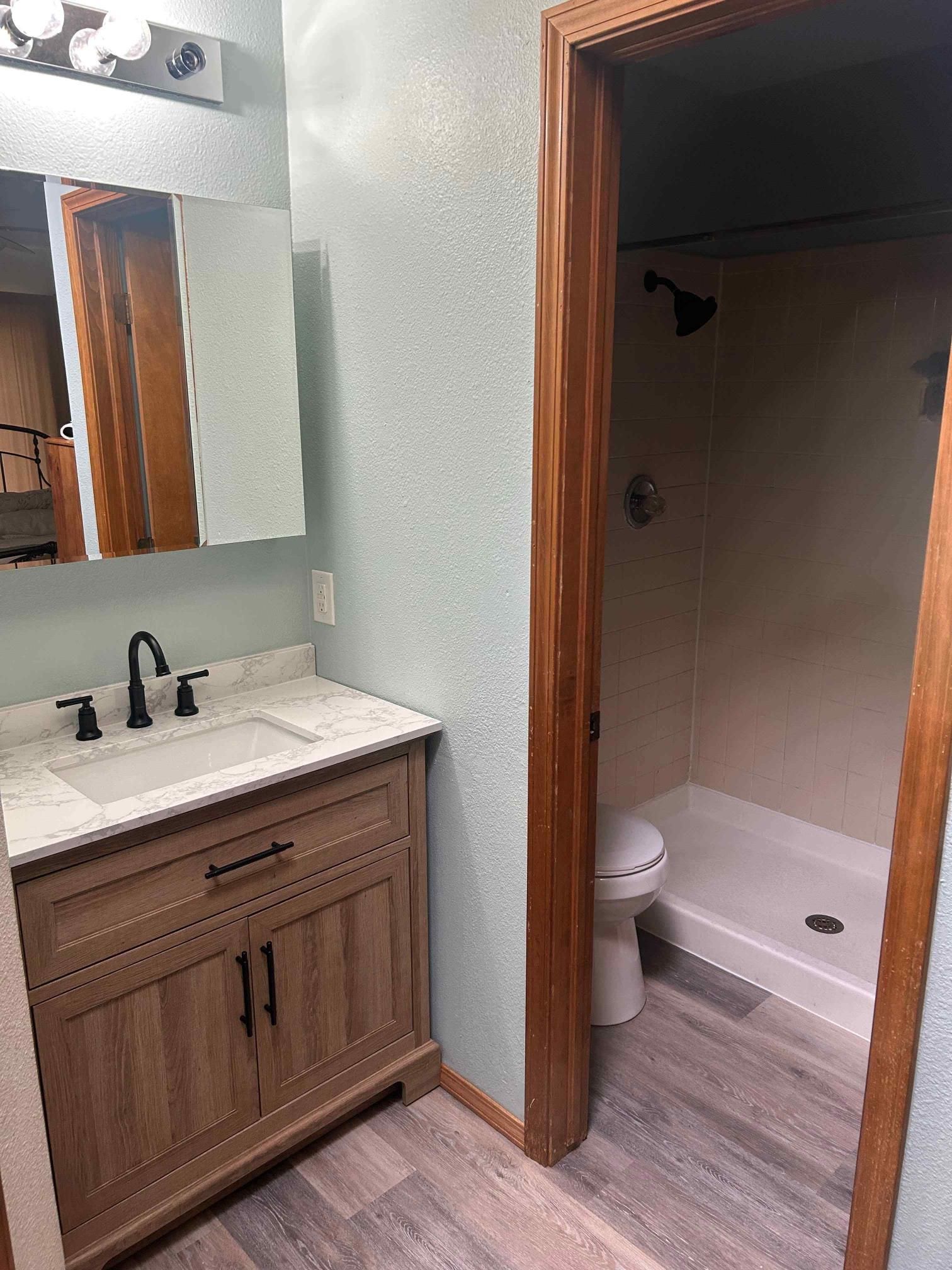 Bathroom with vanity, mirror, and shower. Light blue walls, wood accents, and gray flooring.