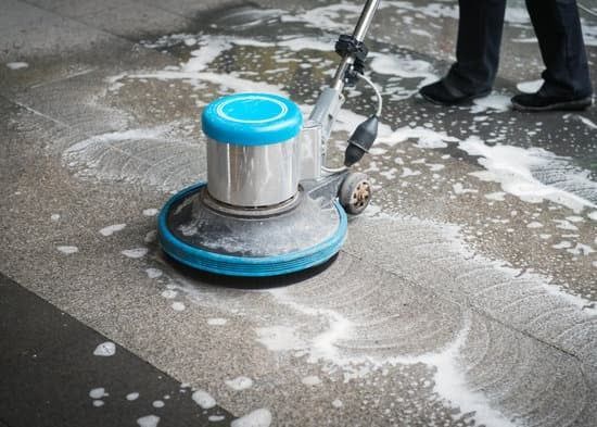 Person using a floor cleaner to scrub a gray concrete surface. Blue and white machine with foamy soap.
