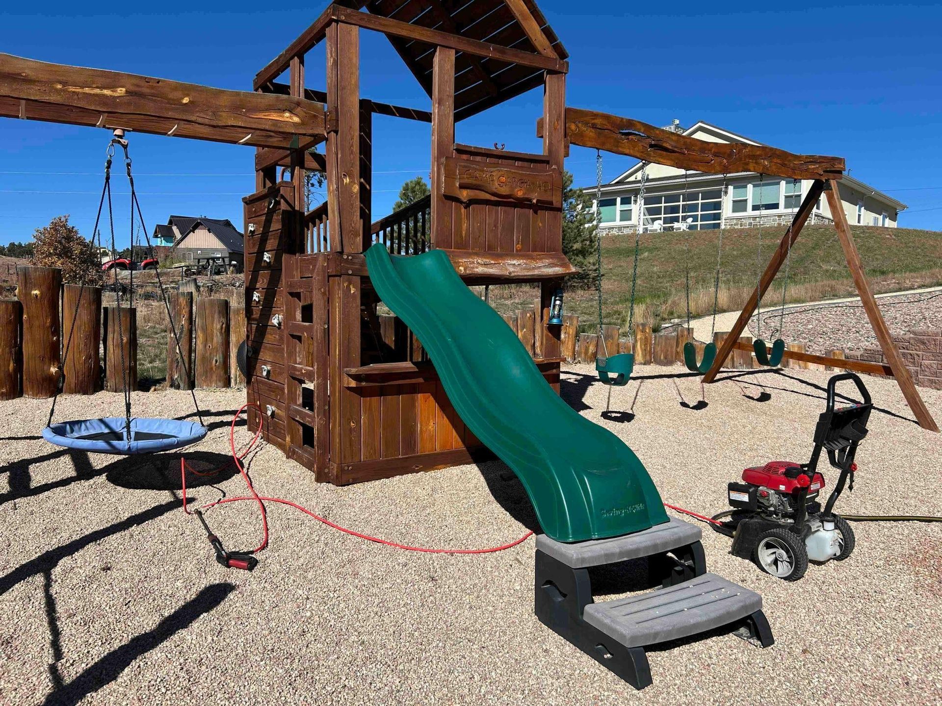 Wooden playset with a green slide and swings on a gravel surface. A pressure washer is nearby.