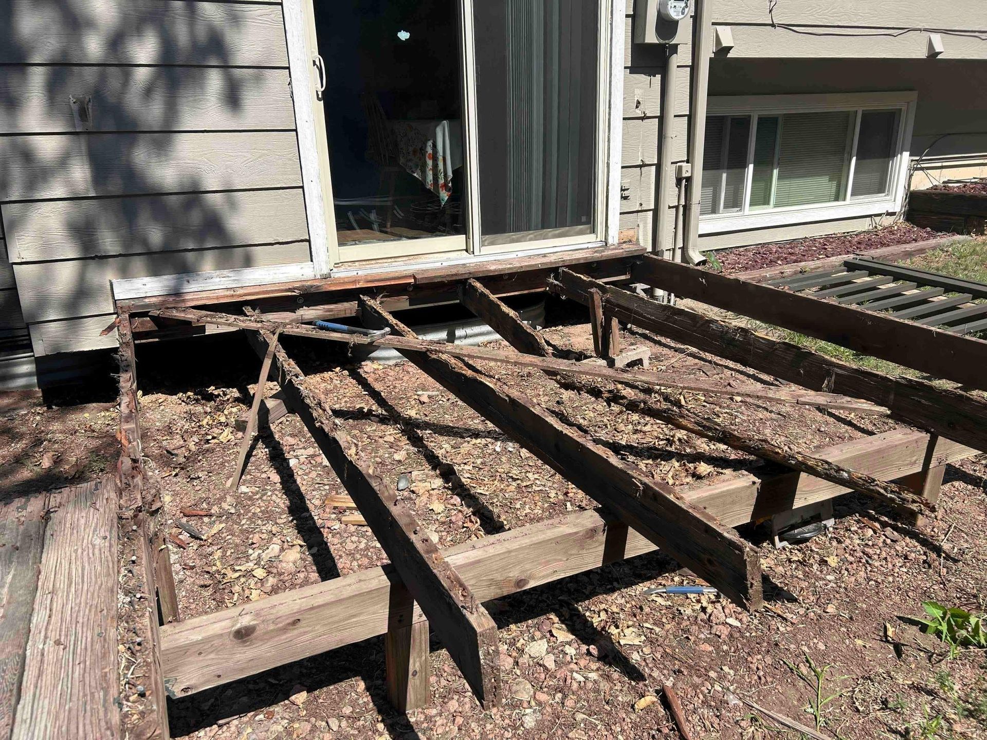 Partially dismantled wooden deck next to a house with a sliding door and window. The wood is weathered and brown.