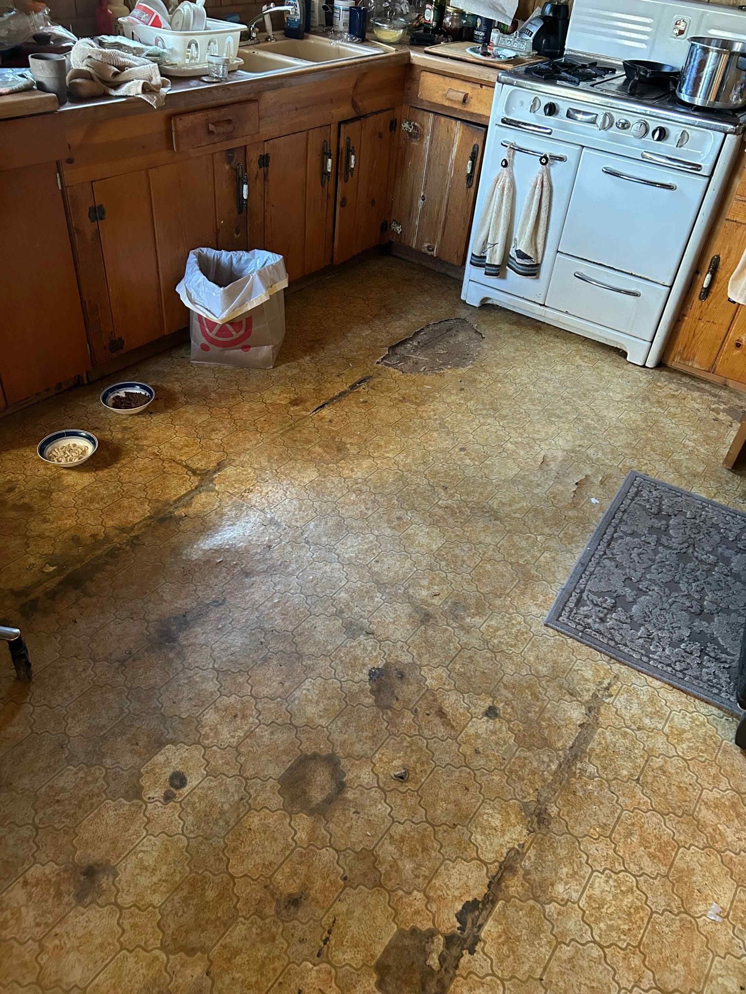 Messy kitchen with cracked linoleum floor, old wooden cabinets, and a stove.