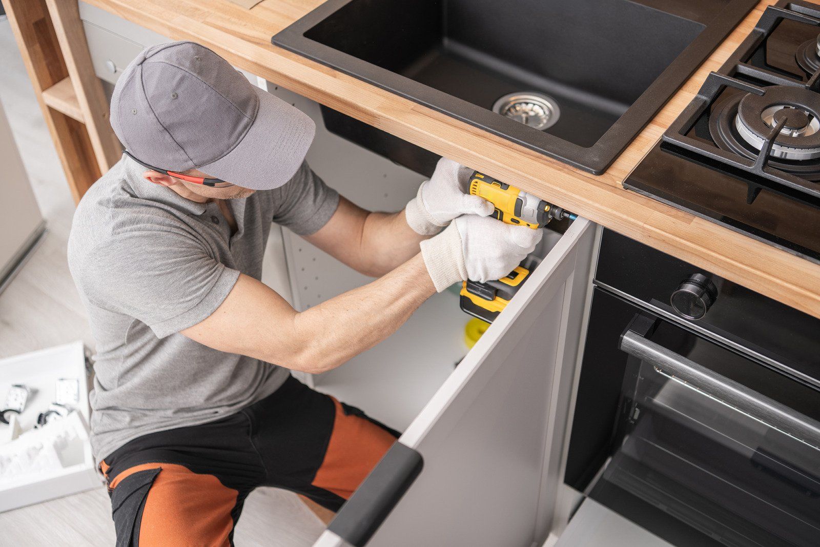 A man installing a cabinet door with a drill under a kitchen sink.