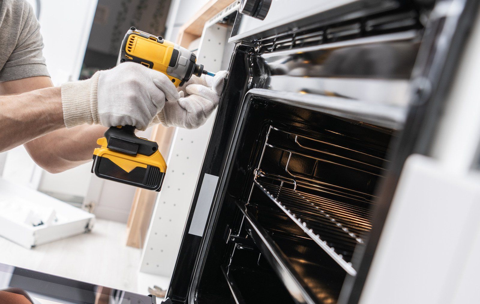 Person wearing gloves uses a power drill to repair an oven in a kitchen.
