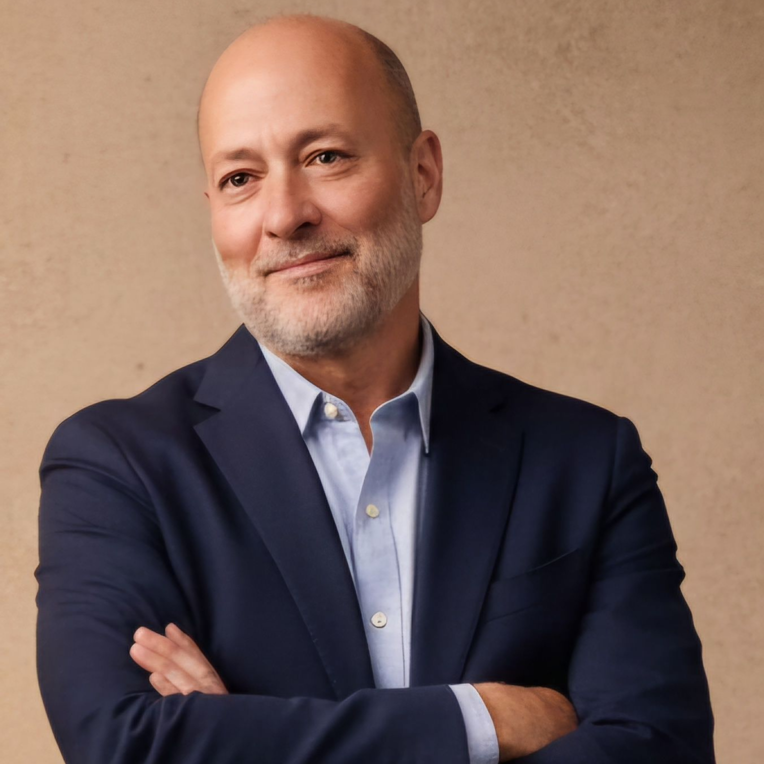 Smiling bald man in a navy blazer with arms crossed against a beige background