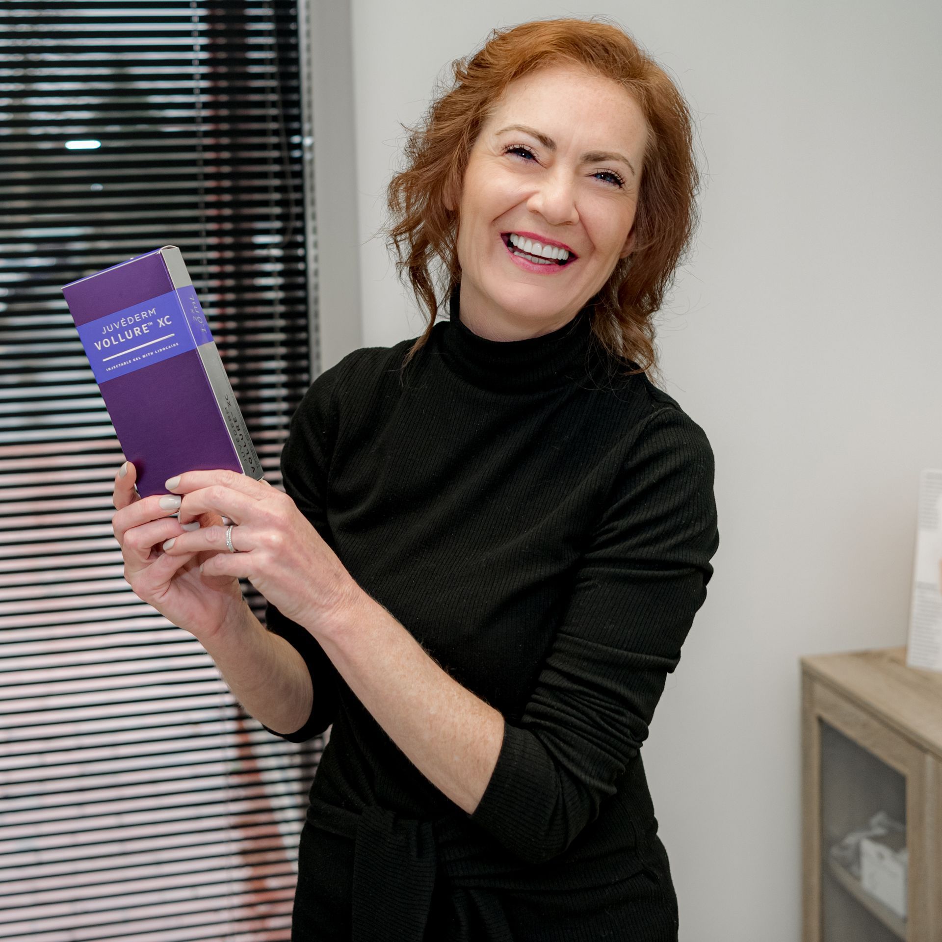 Woman with red hair, smiling, holding a purple box, near a window with blinds.