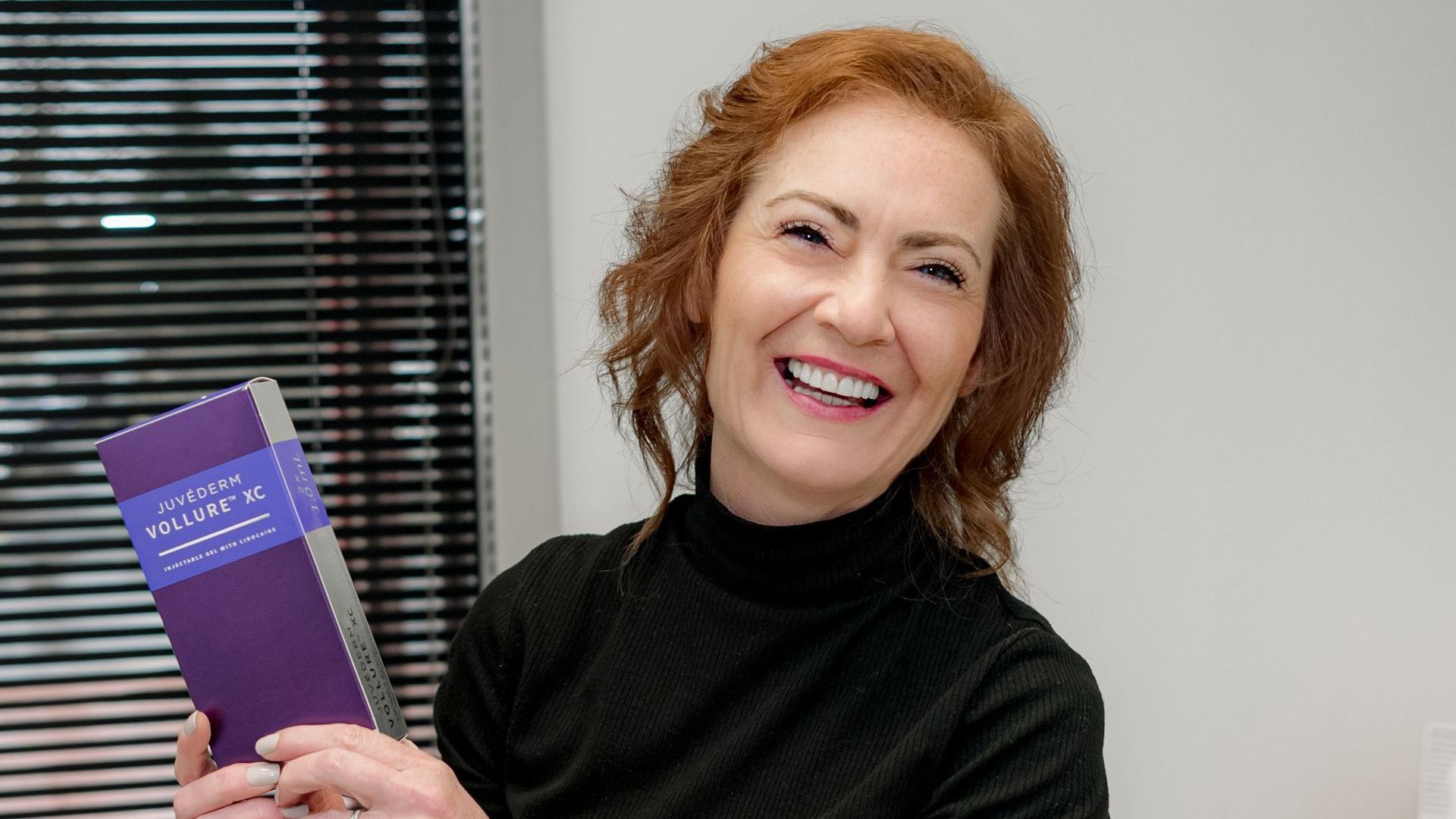 Woman with red hair, smiling, holding a purple book; indoor setting with blinds.