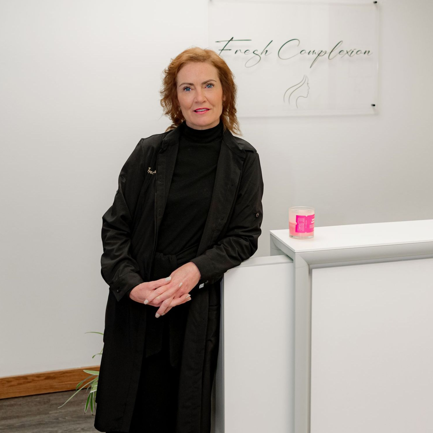 Woman standing beside a white counter in a bright office, wearing a black coat and turtleneck.