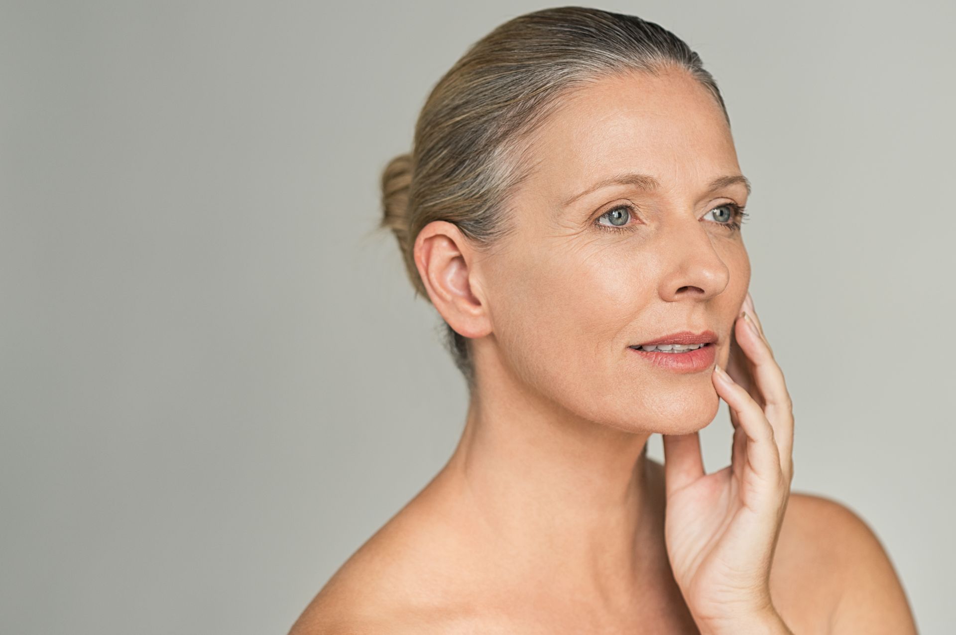 Portrait of a woman with gray hair touching her cheek against a plain gray background