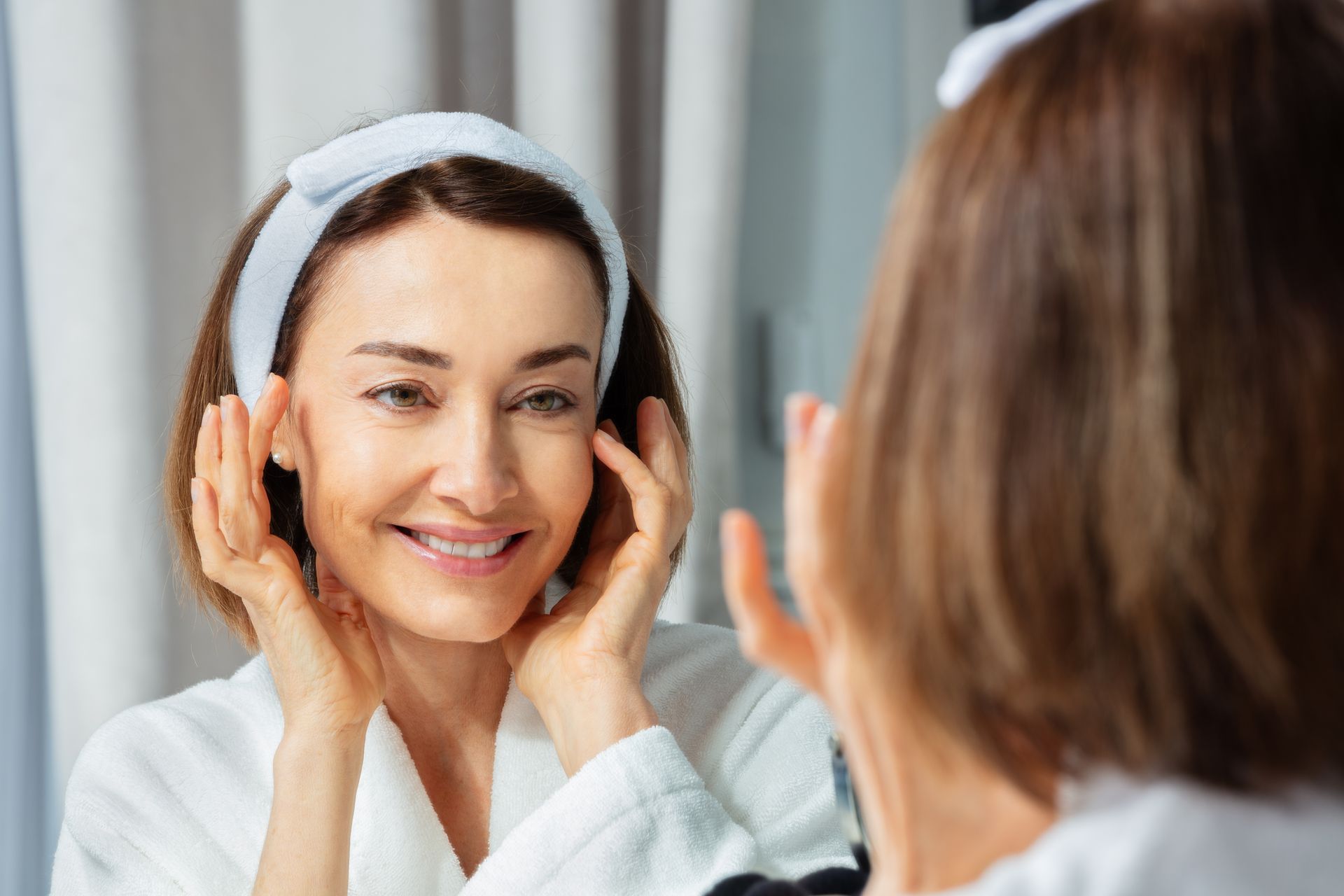 Woman in a white robe applying a facial mask in front of a bathroom mirror