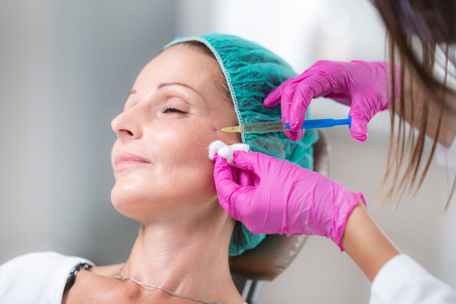 A woman receiving a facial injection while a clinician adjusts her hairnet and holds a syringe.