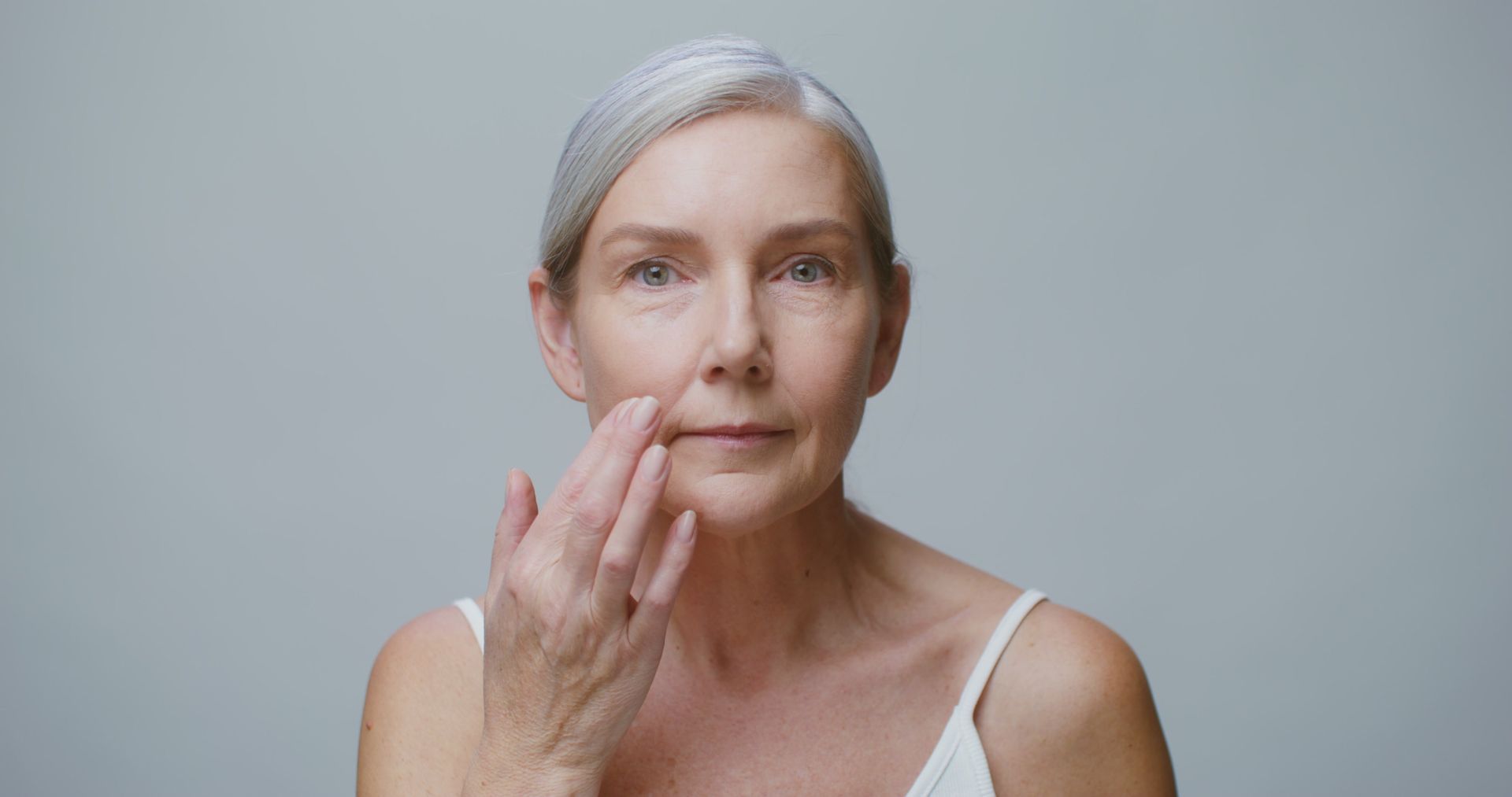 Woman with gray hair touching her face in a soft-lit portrait against a plain background