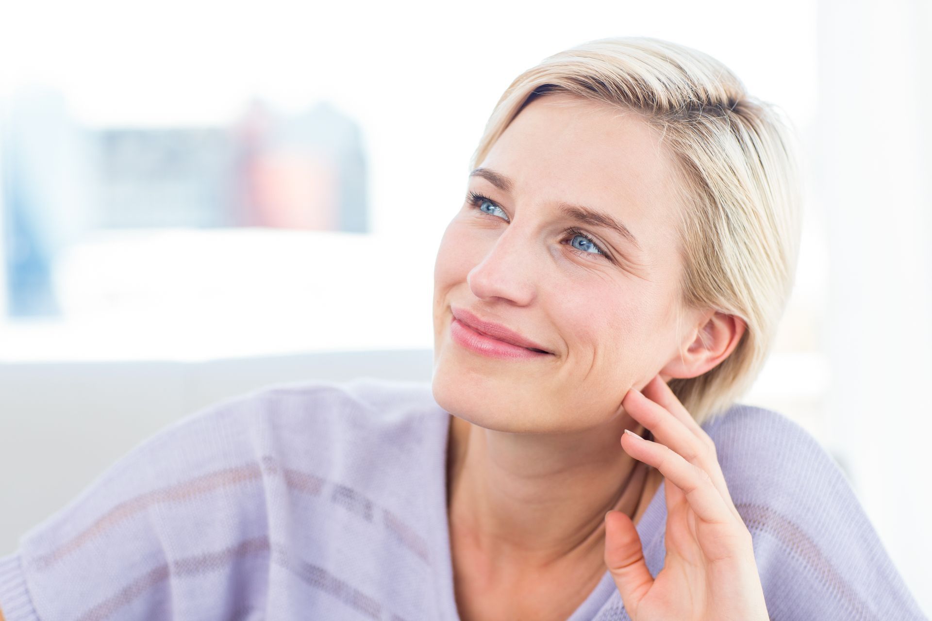 Smiling blonde woman in a light sweater, resting her hand on her cheek against a bright background