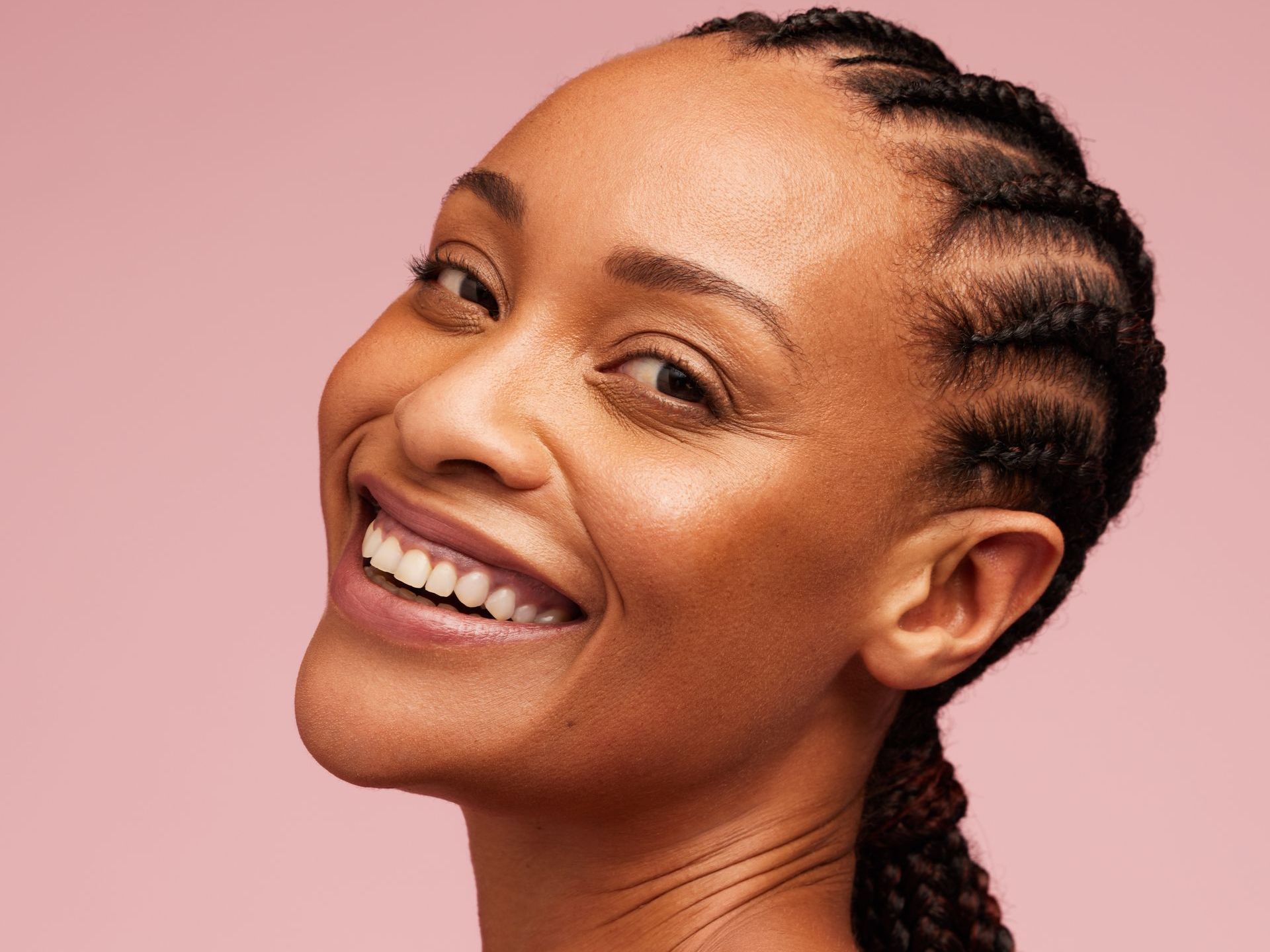 Woman with braided hairstyle smiles broadly, rosy skin, against pink background.