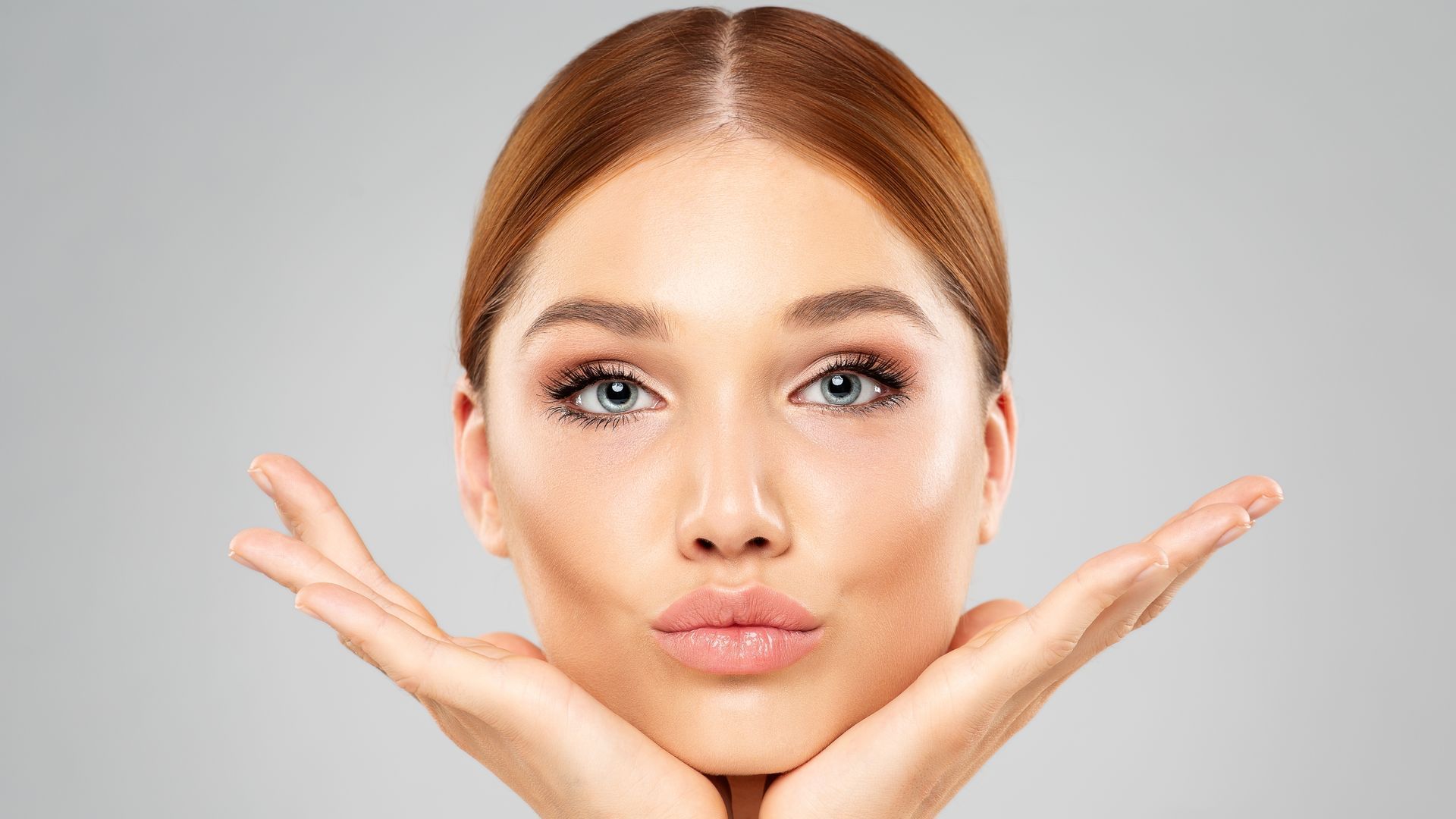 Woman with hands framing her face, puckering lips; light skin, auburn hair, neutral background.